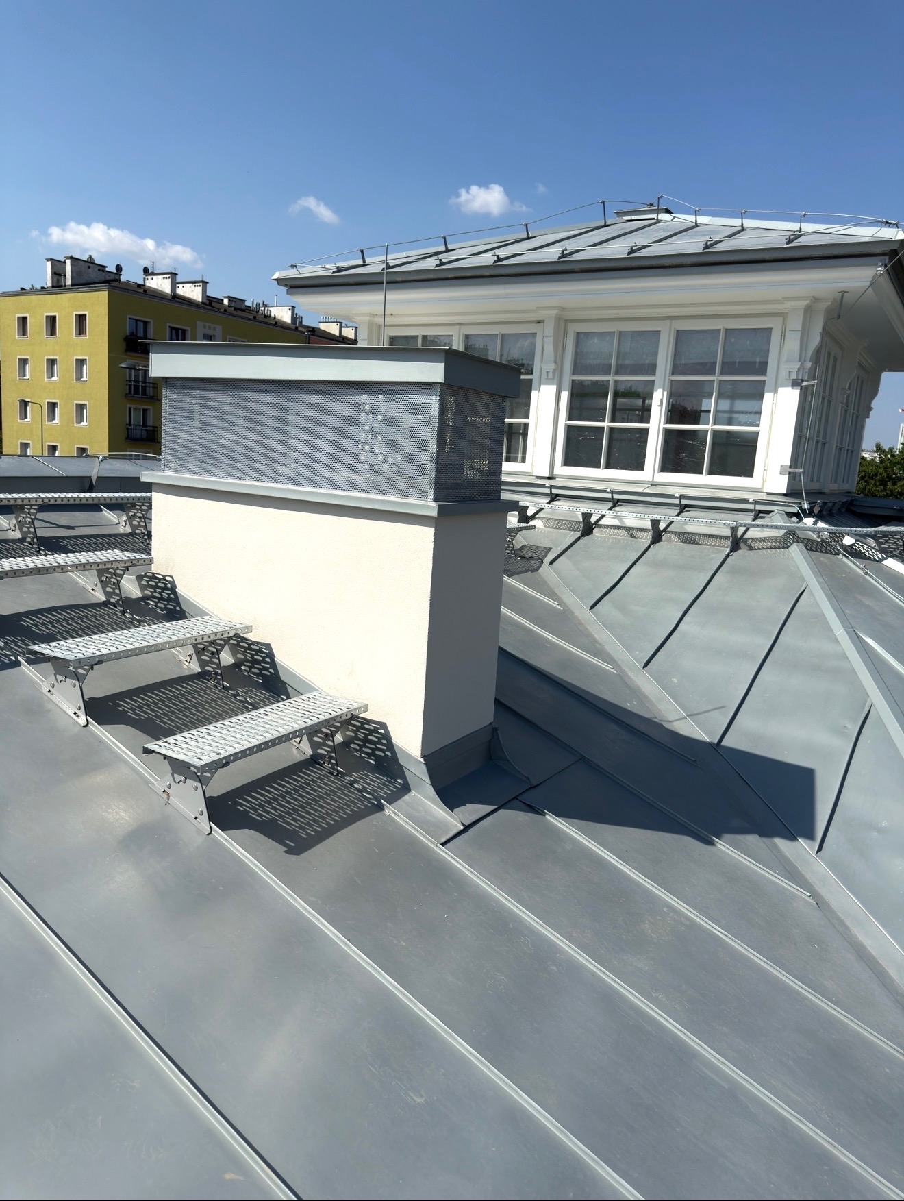 Szary dach z blachy na budynku z kominem i schodkami, widok z góry w słoneczny dzień. W tle budynek mieszkalny i błękitne niebo.