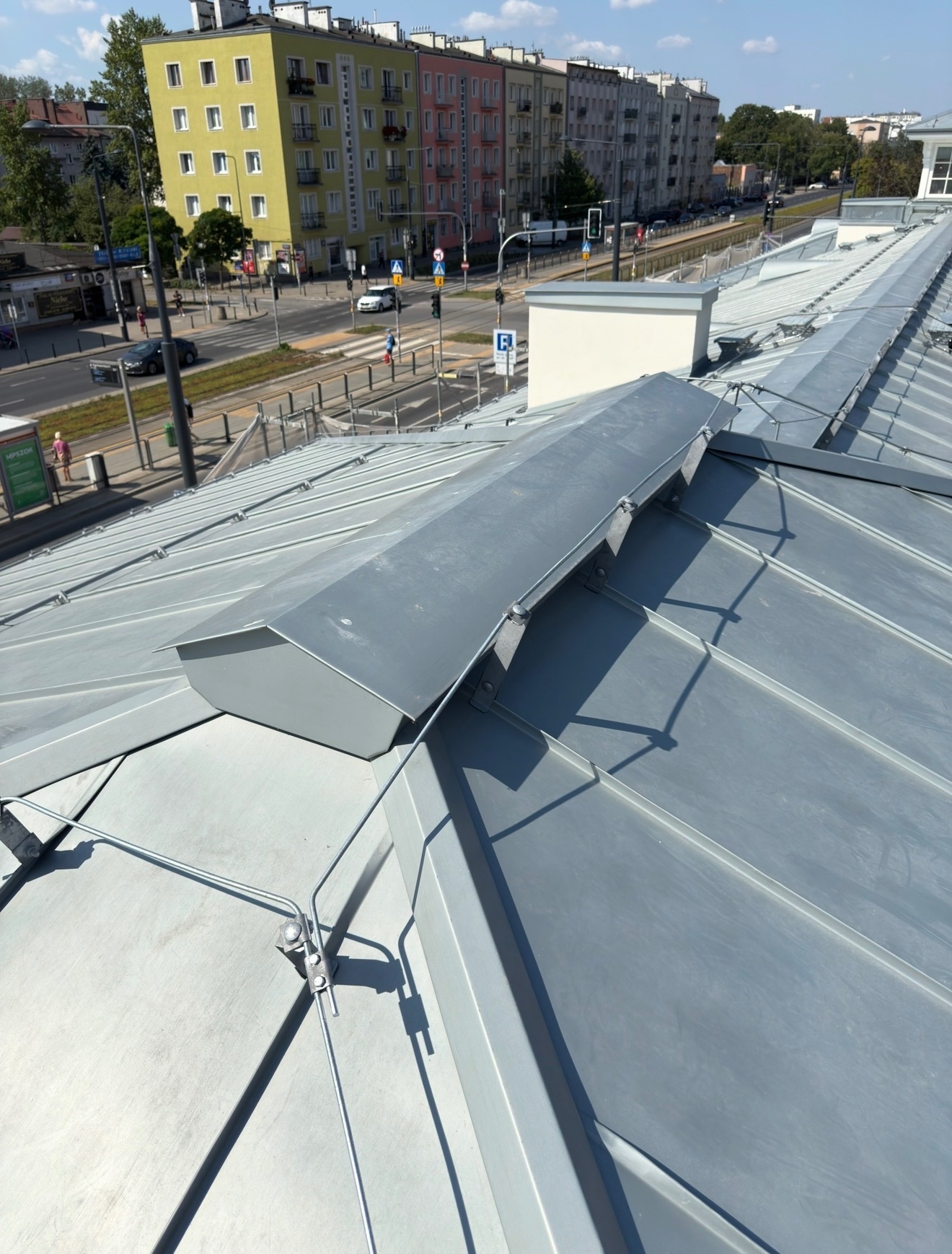 Szary, metalowy dach z widocznymi elementami mocującymi i fragmentem miejskiego krajobrazu w tle, w słoneczny dzień. Widoczne budynki mieszkalne i ulica.