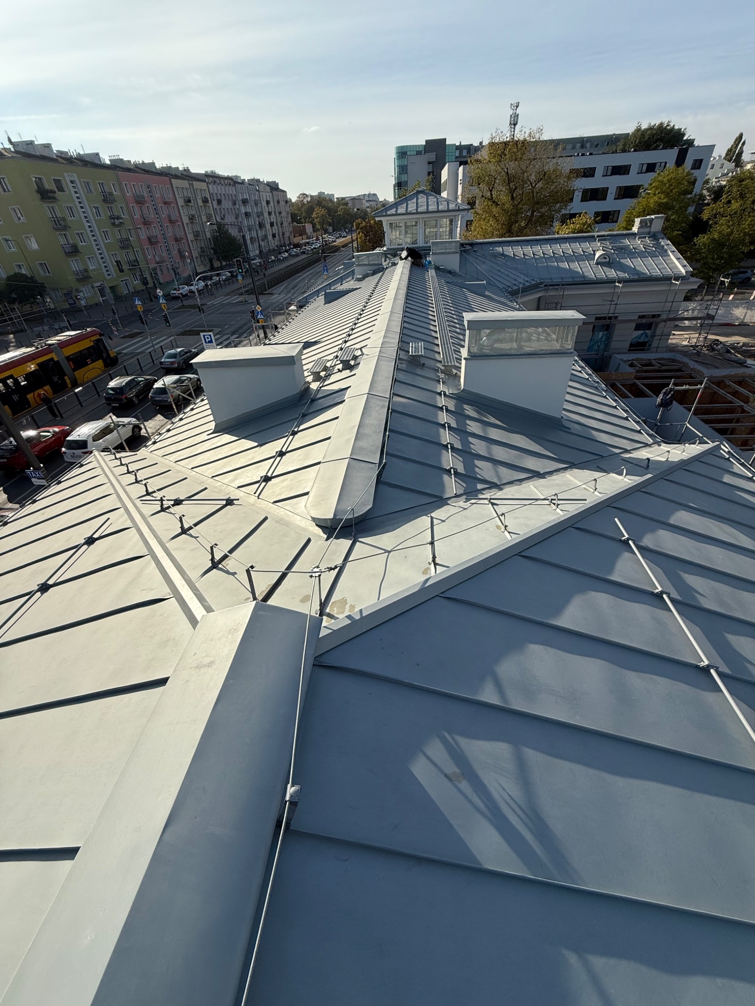 Szary dach z blachy na budynku miejskim, widok z góry. Elementy wentylacyjne i zabezpieczenia przeciwśniegowe. W tle zabudowa miejska i tramwaj.