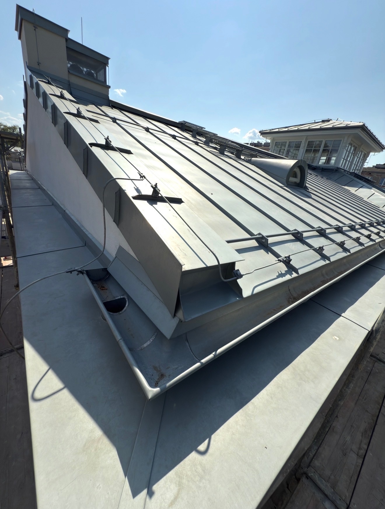 Szary, metalowy dach z widocznymi obróbkami blacharskimi i elementami mocującymi, sfotografowany pod kątem, z fragmentem lukarny w tle i błękitnym niebem.