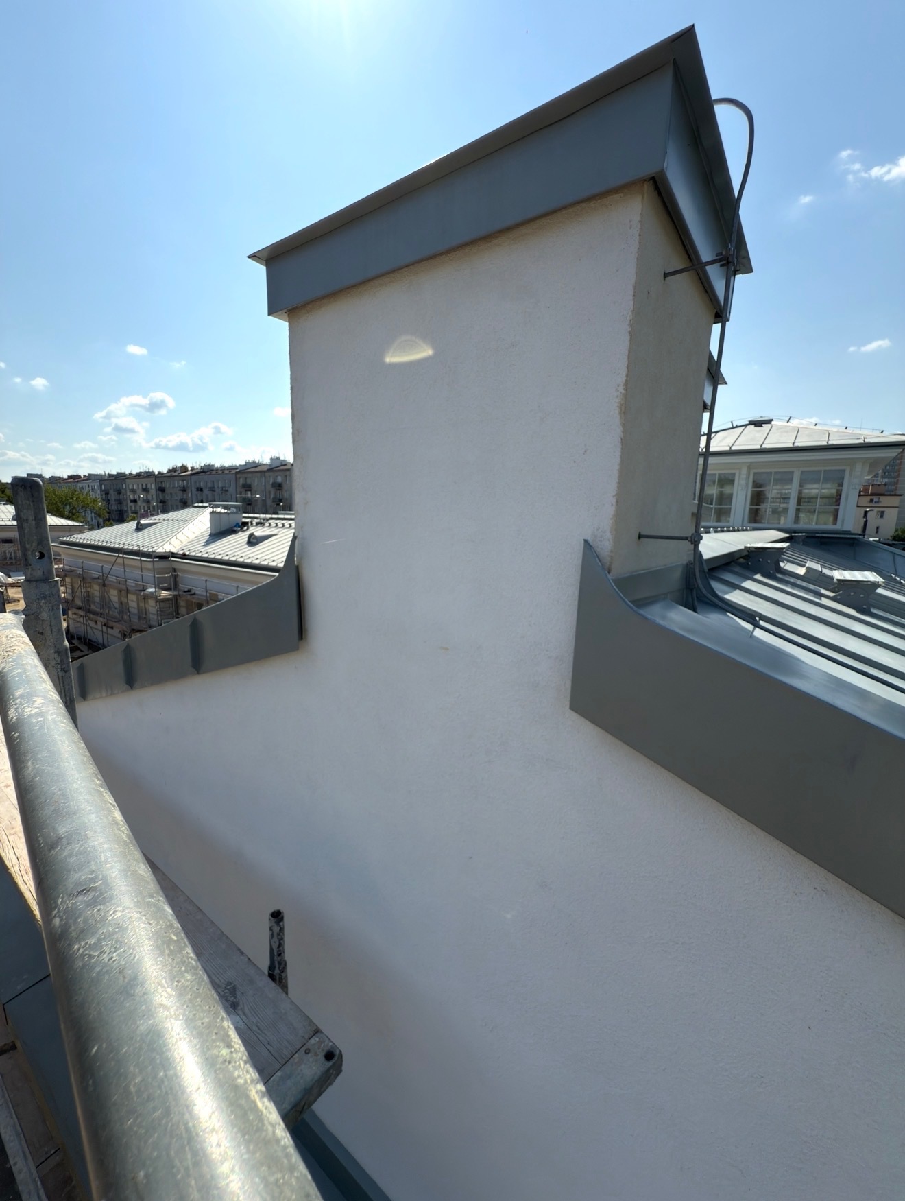 Biały komin z szarą obróbką blacharską na dachu budynku, widok z rusztowania w słoneczny dzień. Detal wykończenia dachu z blachy.