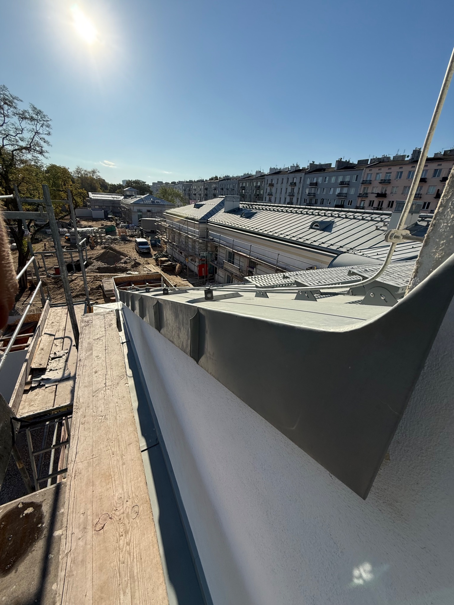 Szary element obróbki blacharskiej na dachu budynku w trakcie prac. Widoczne rusztowania, budynki w tle i błękitne niebo. Ujęcie z góry, w słoneczny dzień.
