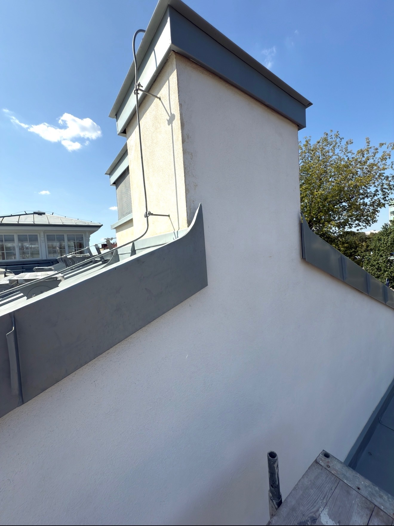 Szczegółowe ujęcie komina z obróbkami blacharskimi na dachu budynku, widoczne elementy konstrukcyjne i drabinka kominiarska na tle błękitnego nieba.