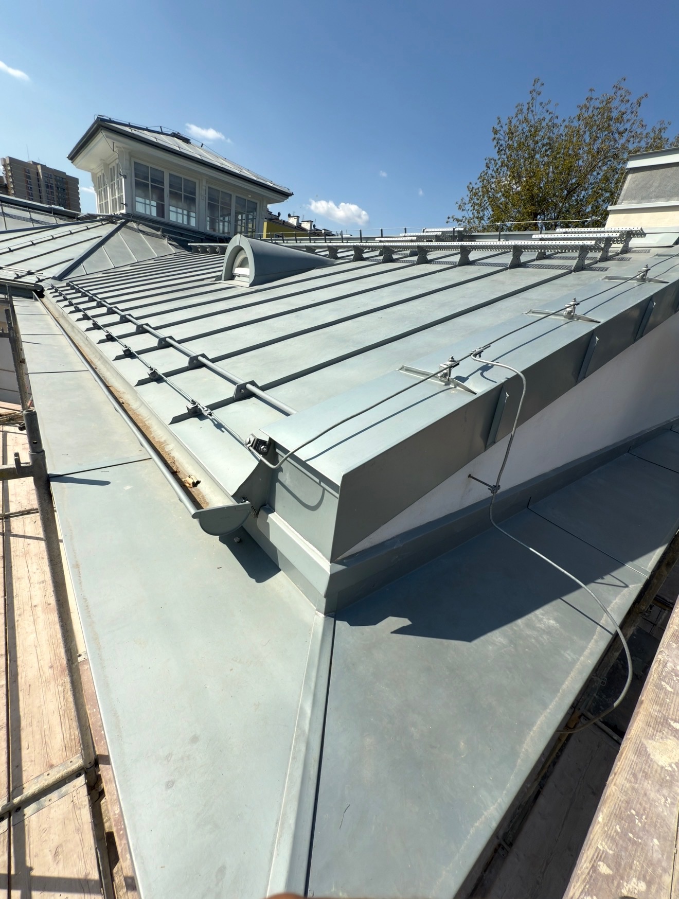 Szary dach z blachy na budynku z lukarną, widoczny z rusztowania. Metalowe obróbki i mocowania na dachu, błękitne niebo w tle. Ujęcie z dołu, podkreślające detale.