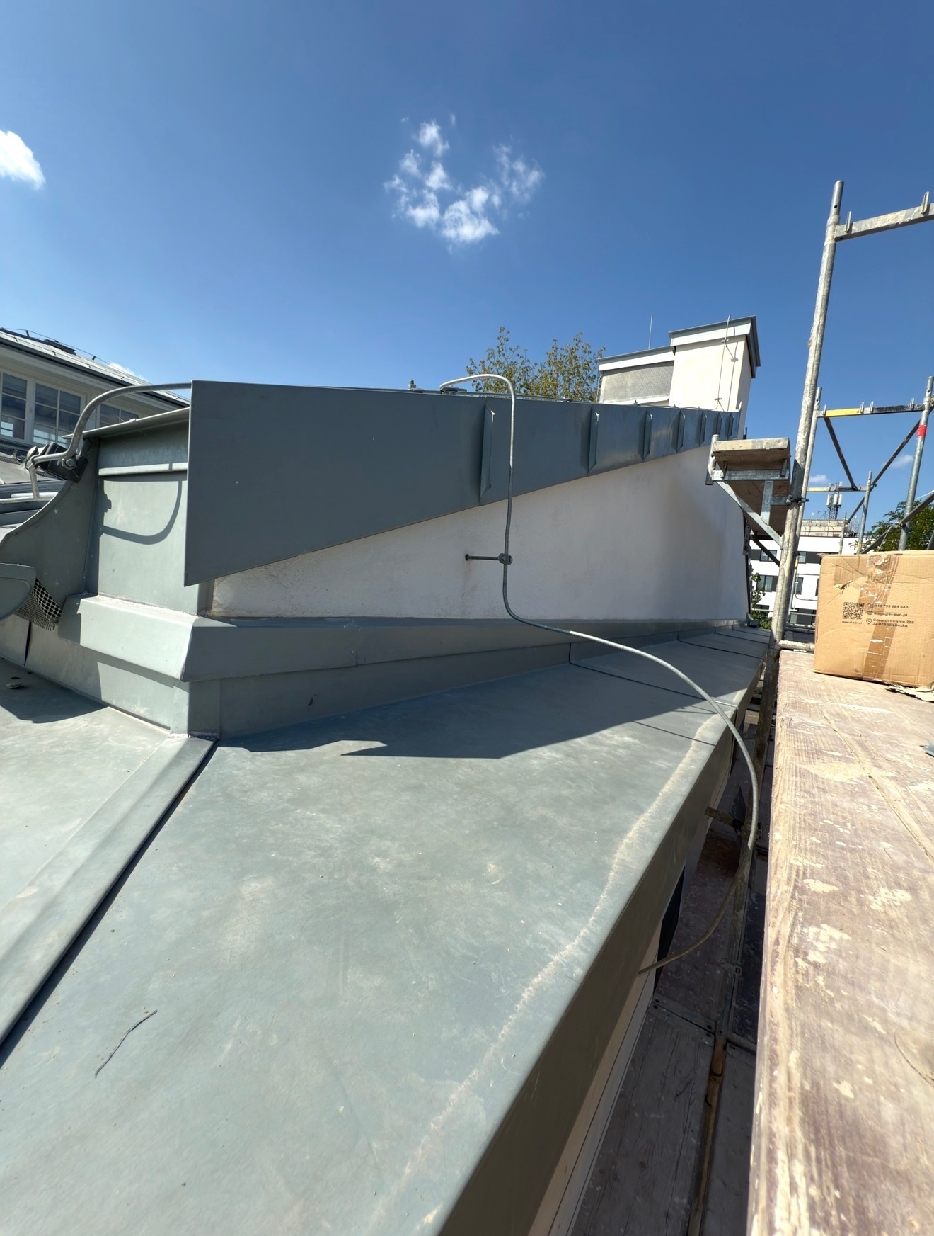 Szary, płaski dach z obróbkami blacharskimi, widok z góry, fragment rusztowania i karton na dachu, błękitne niebo w tle. Detal wykonania.