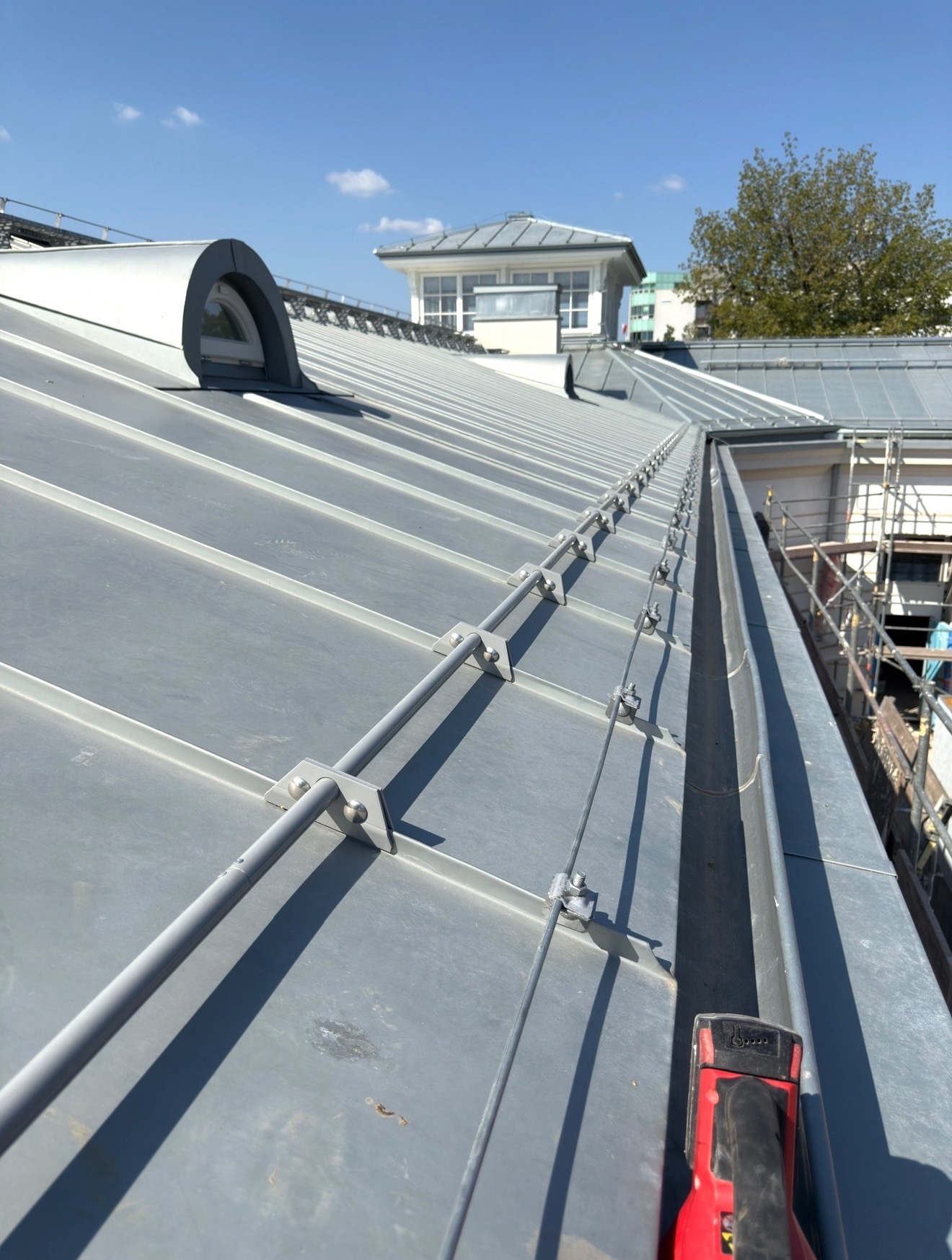 Szary dach z blachy z systemem odgromowym i lukarną. Widoczny fragment rynny i narzędzie dekarskie. W tle budynek z wieżyczką i drzewo na tle błękitnego nieba.