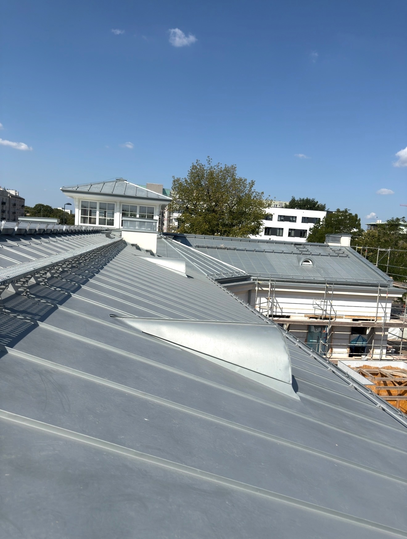 Szary dach z blachy na budynku z lukarną, widoczne zabezpieczenia przeciwśniegowe i rusztowania. Błękitne niebo w tle.