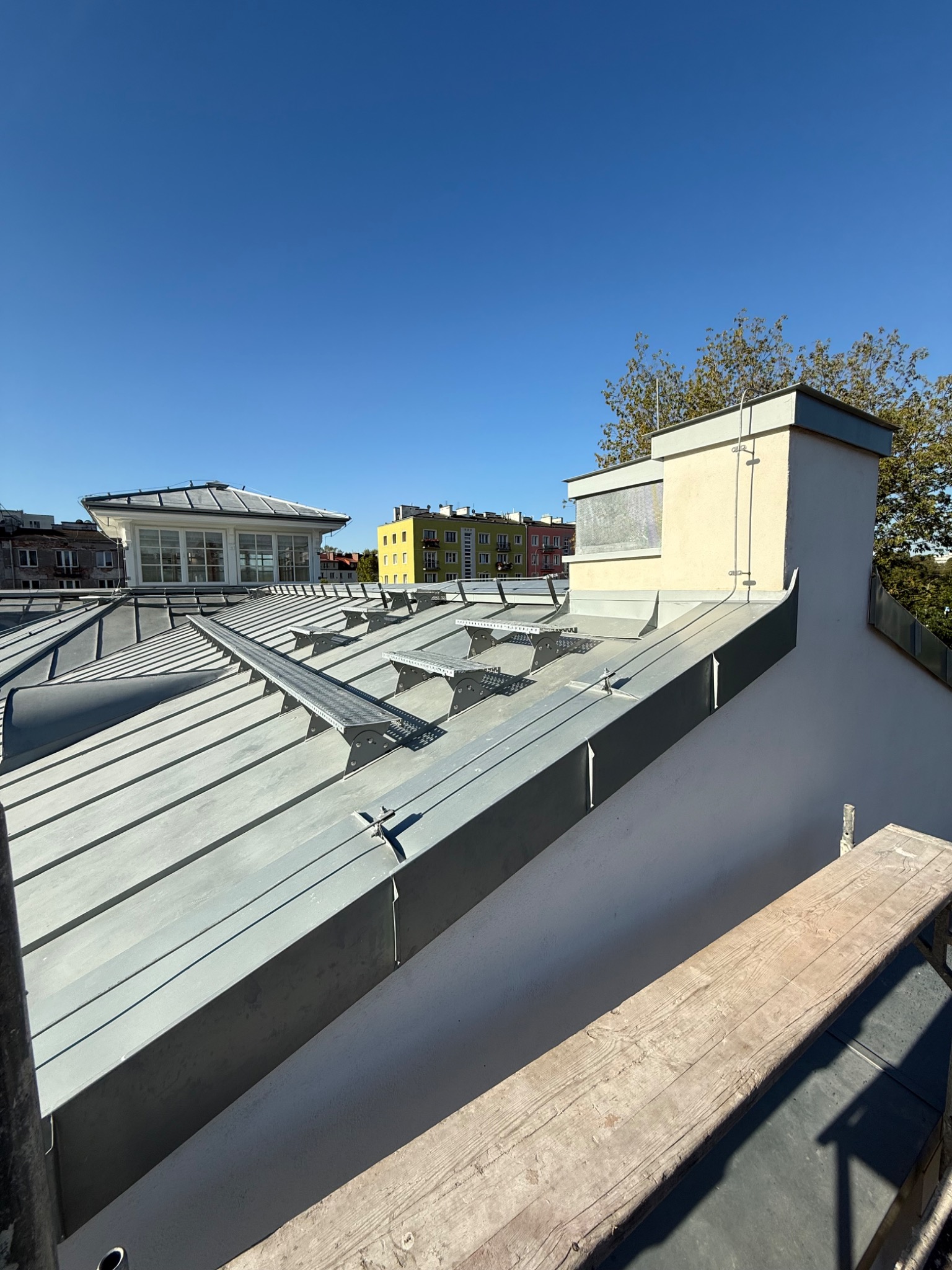 Szary dach z blachy na budynku z kominem, widoczne elementy mocujące i fragment rusztowania. W tle budynki mieszkalne i błękitne niebo.
