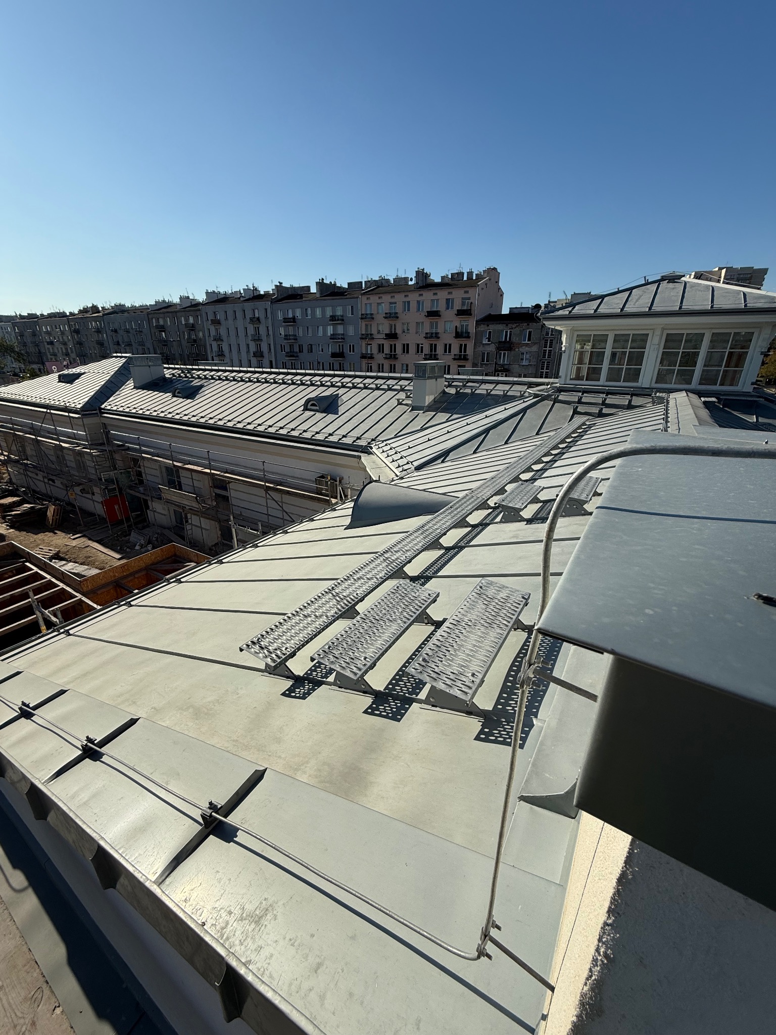 Szary dach z blachy z elementami bezpieczeństwa, widok z góry na tle zabudowy miejskiej i błękitnego nieba. Widoczne metalowe stopnie i zabezpieczenia przeciwśniegowe.