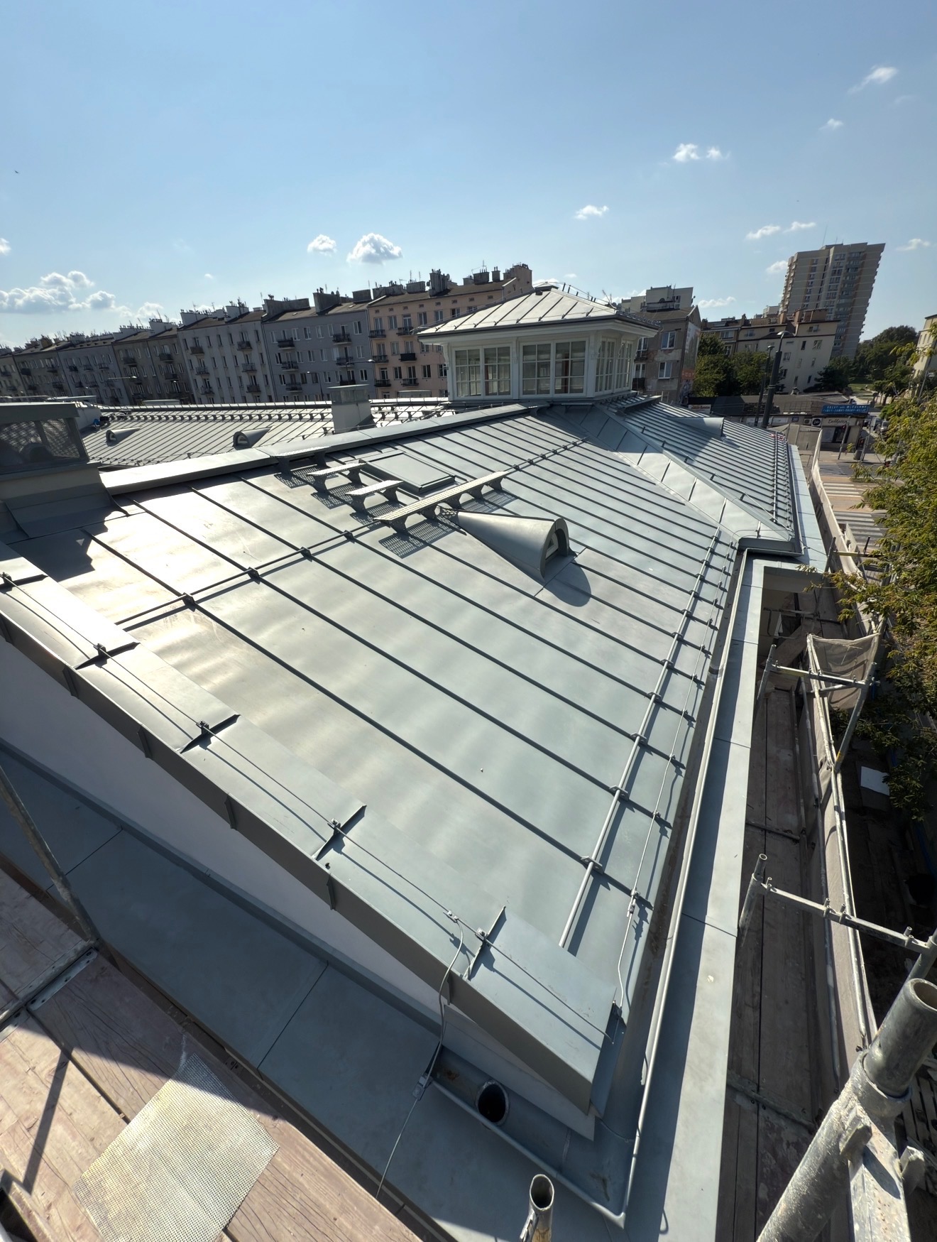 Szary, metalowy dach z widocznymi obróbkami blacharskimi i elementami wentylacyjnymi na tle miejskiej zabudowy w słoneczny dzień. Widoczne rusztowania.