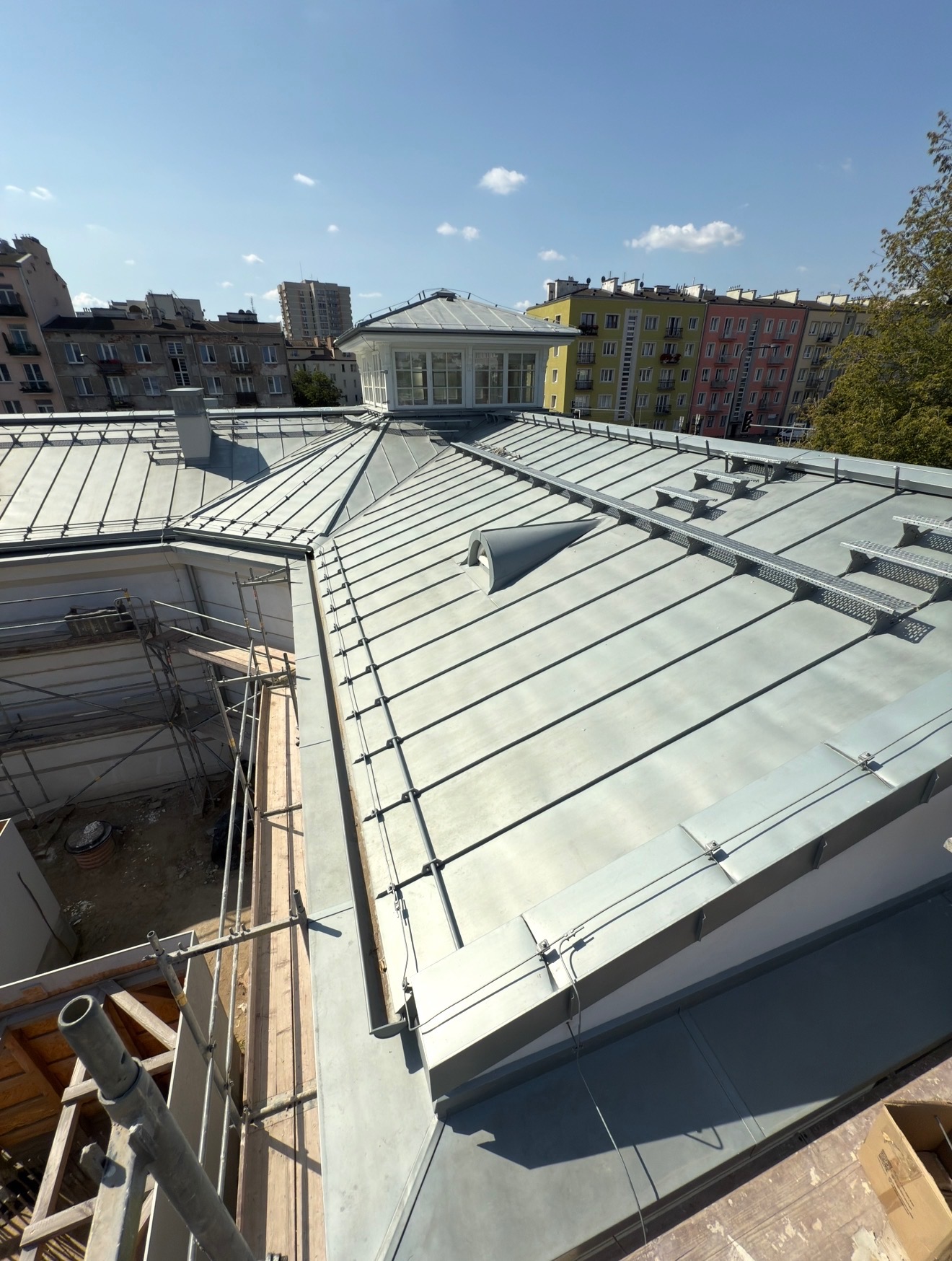 Metalowy dach z lukarną na tle miejskiej zabudowy w słoneczny dzień. Widoczne elementy konstrukcyjne i rusztowania, sugerujące etap prac budowlanych lub remontowych.