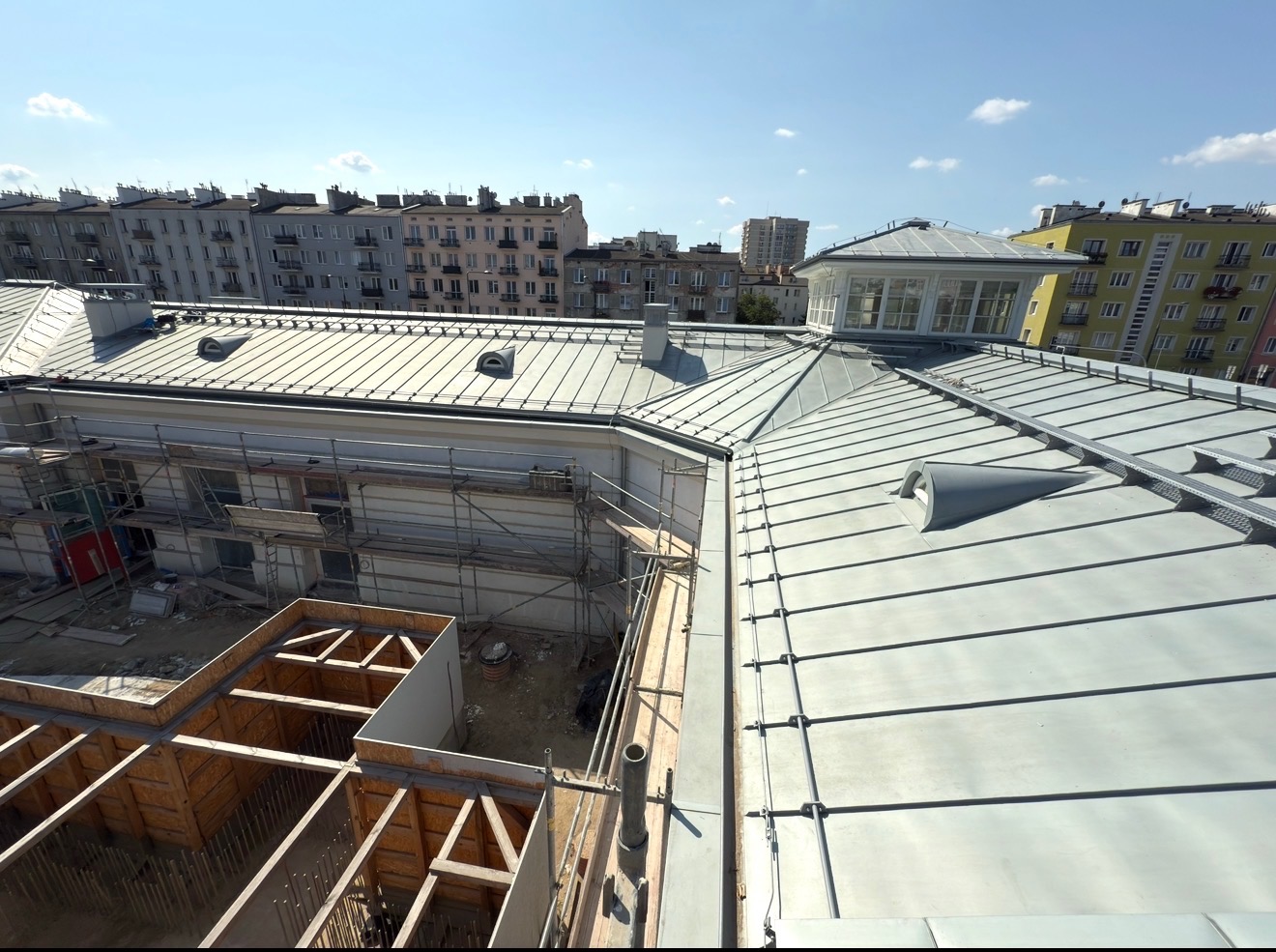 Metalowy dach z lukarnami w trakcie prac, widok z góry na budynek w rusztowaniach i konstrukcję tarasu. W tle zabudowa miejska i błękitne niebo.