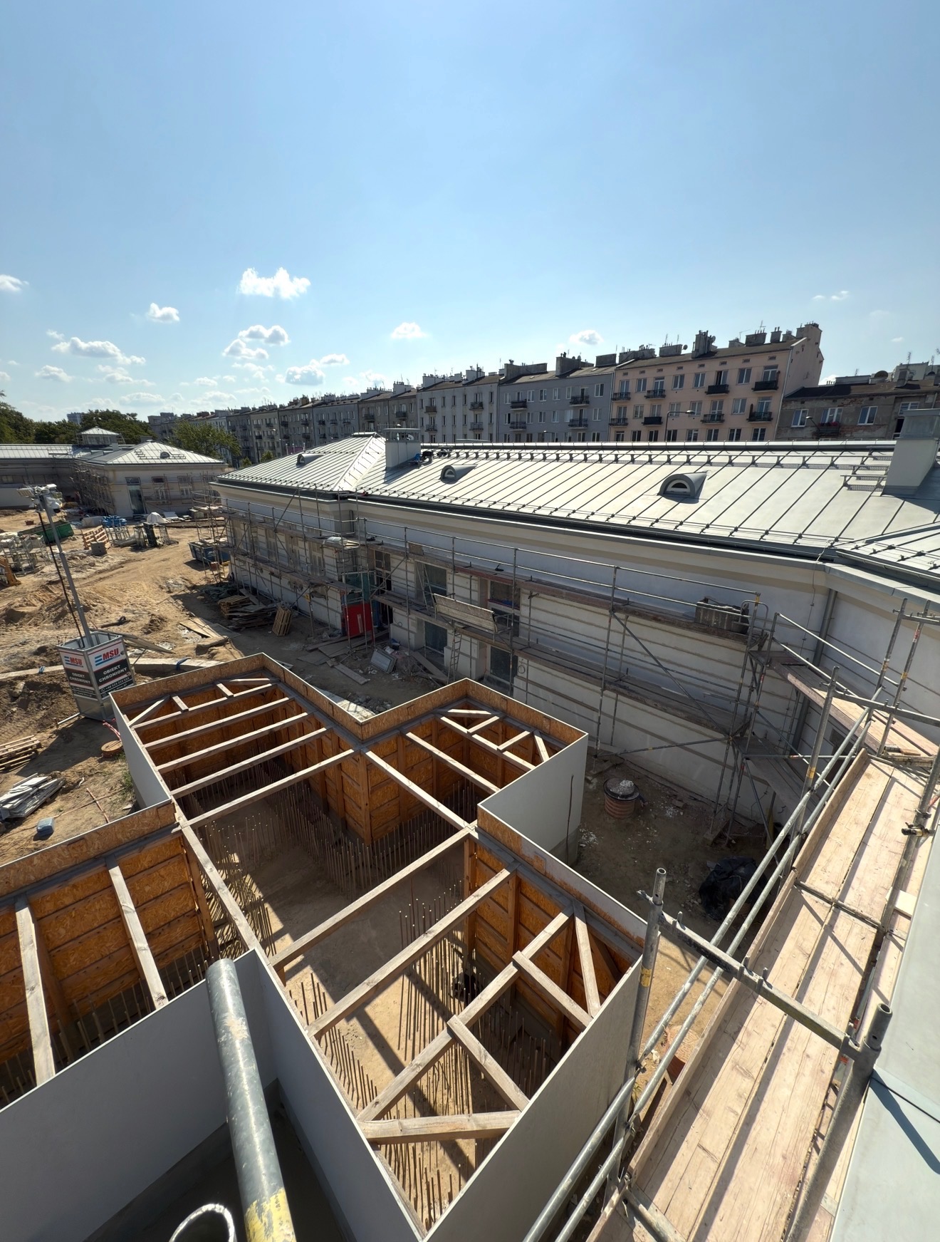 Budowa budynku z widocznym szkieletem konstrukcyjnym i rusztowaniami. W tle budynki mieszkalne pod błękitnym niebem z chmurami.