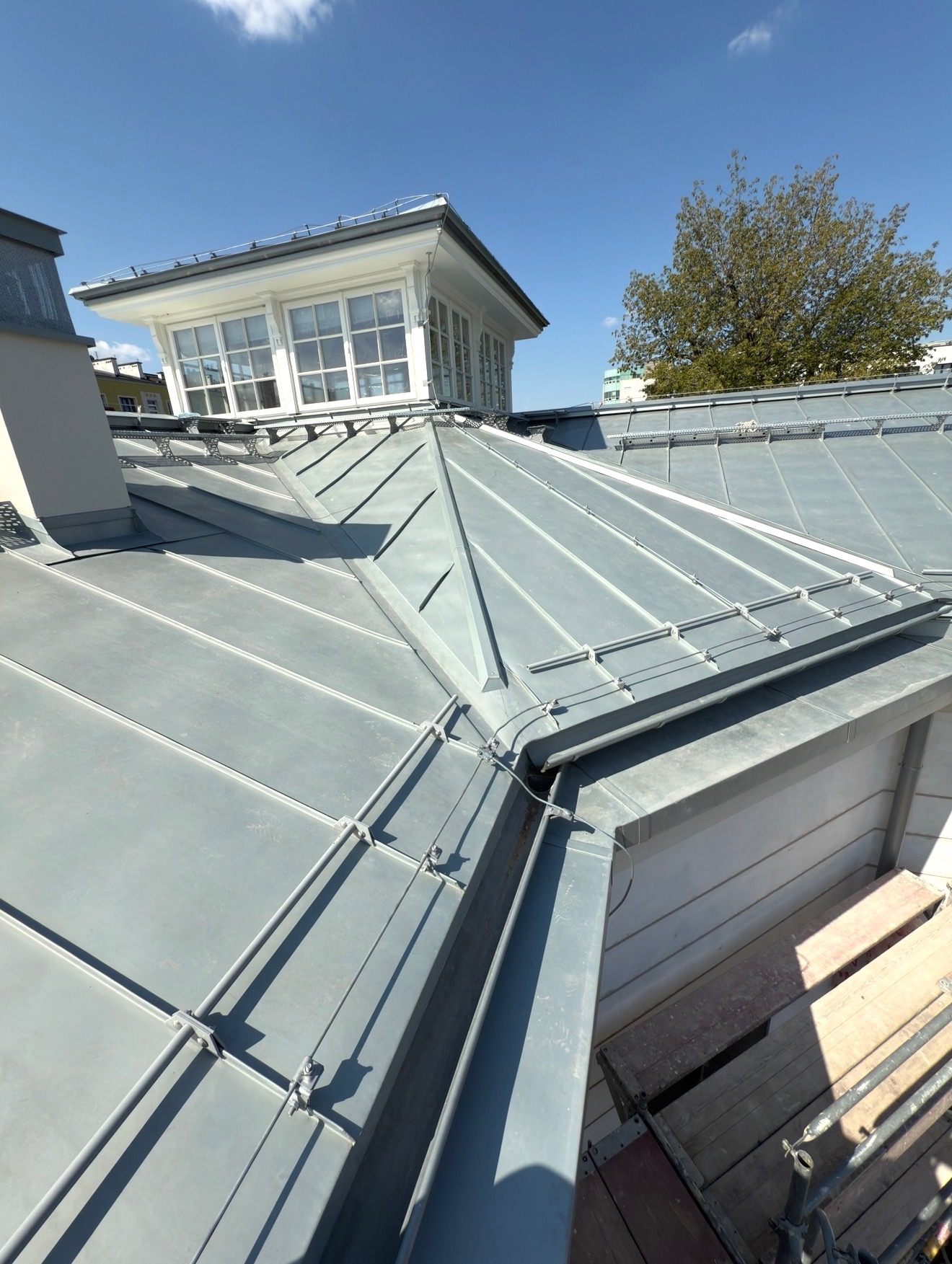 Szary dach z blachy z lukarną i systemem odgromowym, widoczny fragment rusztowania. Ujęcie z góry, w tle błękitne niebo i drzewo.