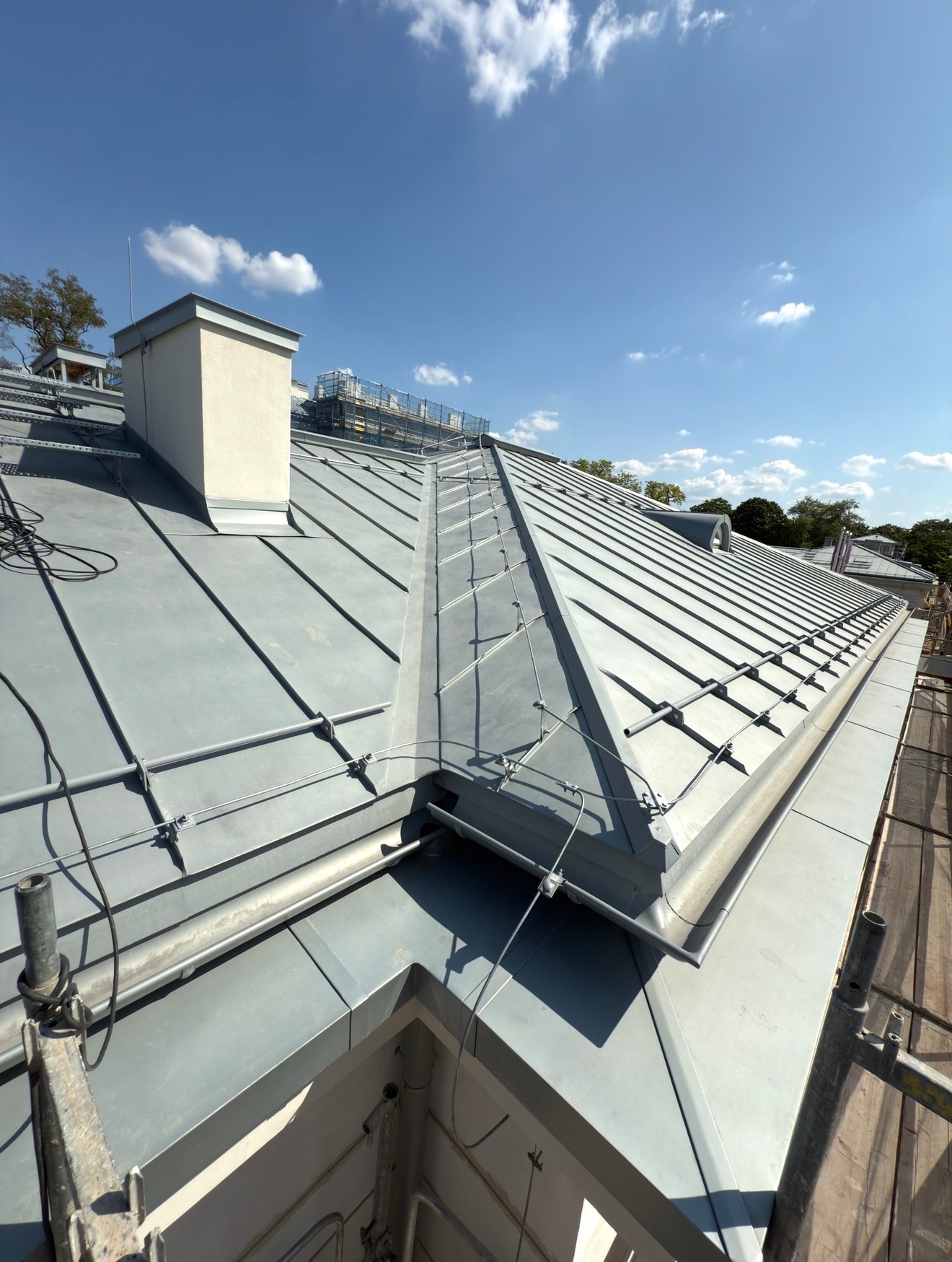 Szary dach z blachy na budynku w słoneczny dzień. Widoczne obróbki blacharskie, komin i fragment rusztowania. Dach pokryty blachą na rąbek stojący.