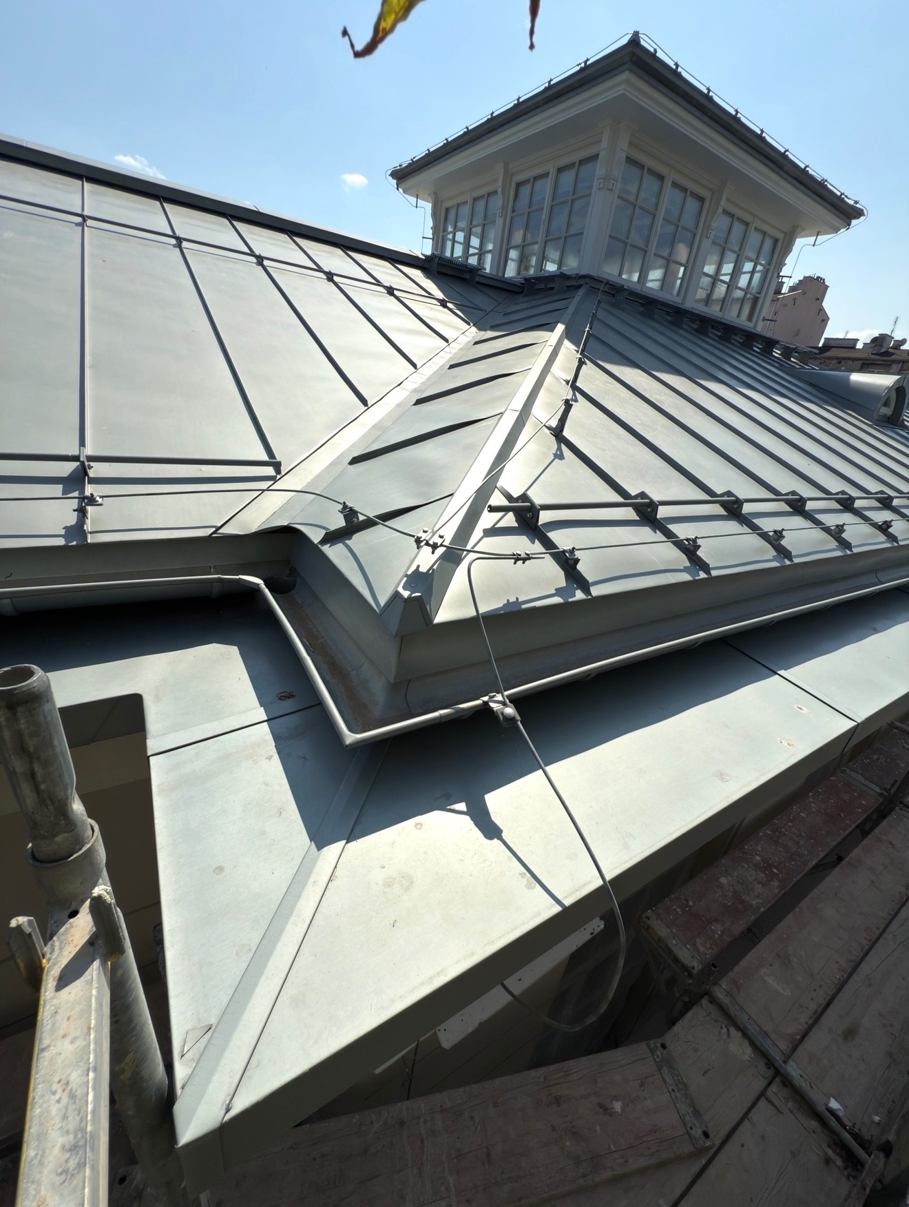 Szary dach z blachy na budynku w Warszawie, widoczna lukarna i fragment rusztowania. Metalowe obróbki blacharskie i zabezpieczenia przeciwśniegowe.