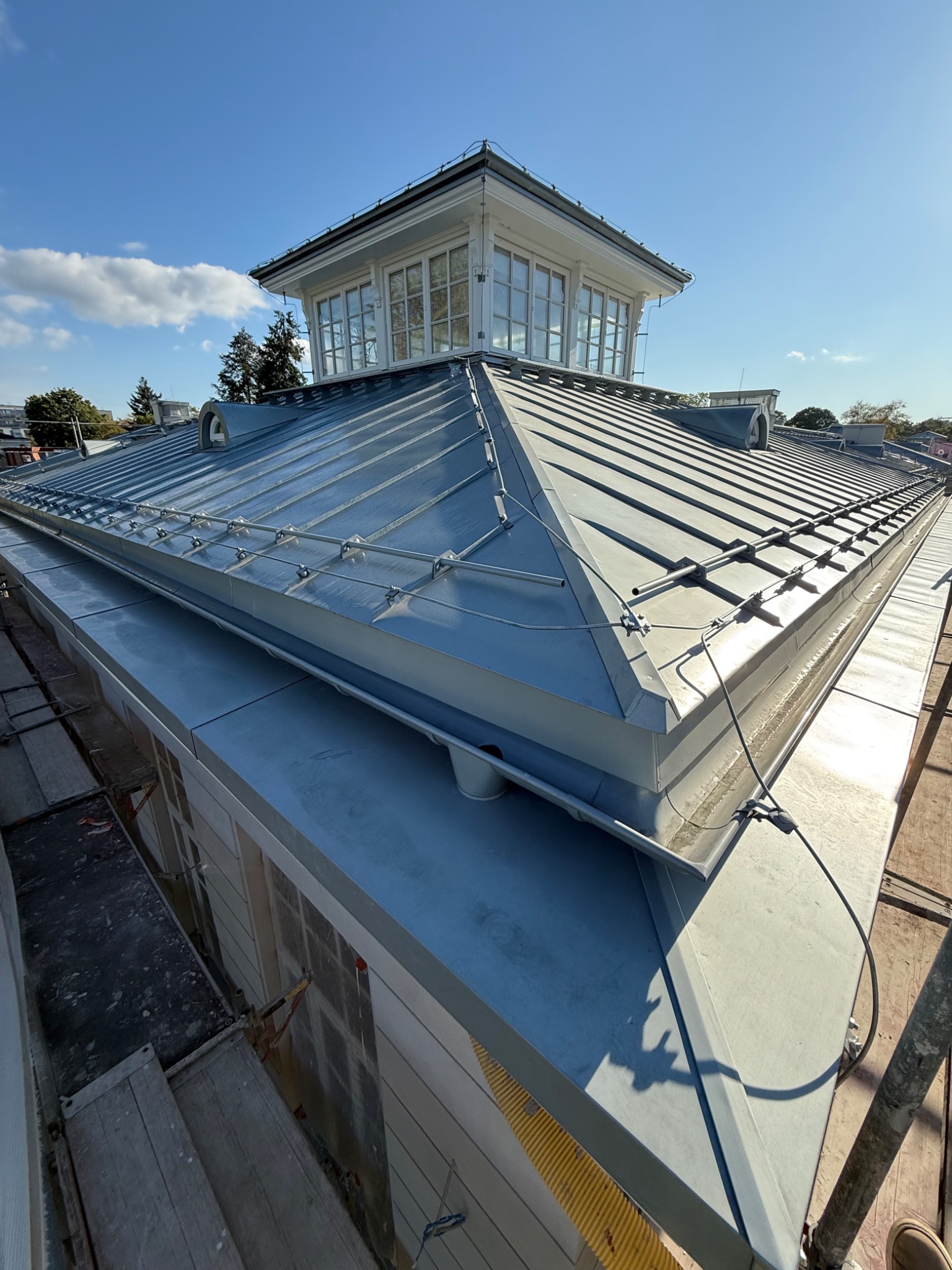 Szary dach z blachy z lukarną i systemem odgromowym, widoczny z rusztowania w słoneczny dzień. Detal obróbek blacharskich i mocowań.