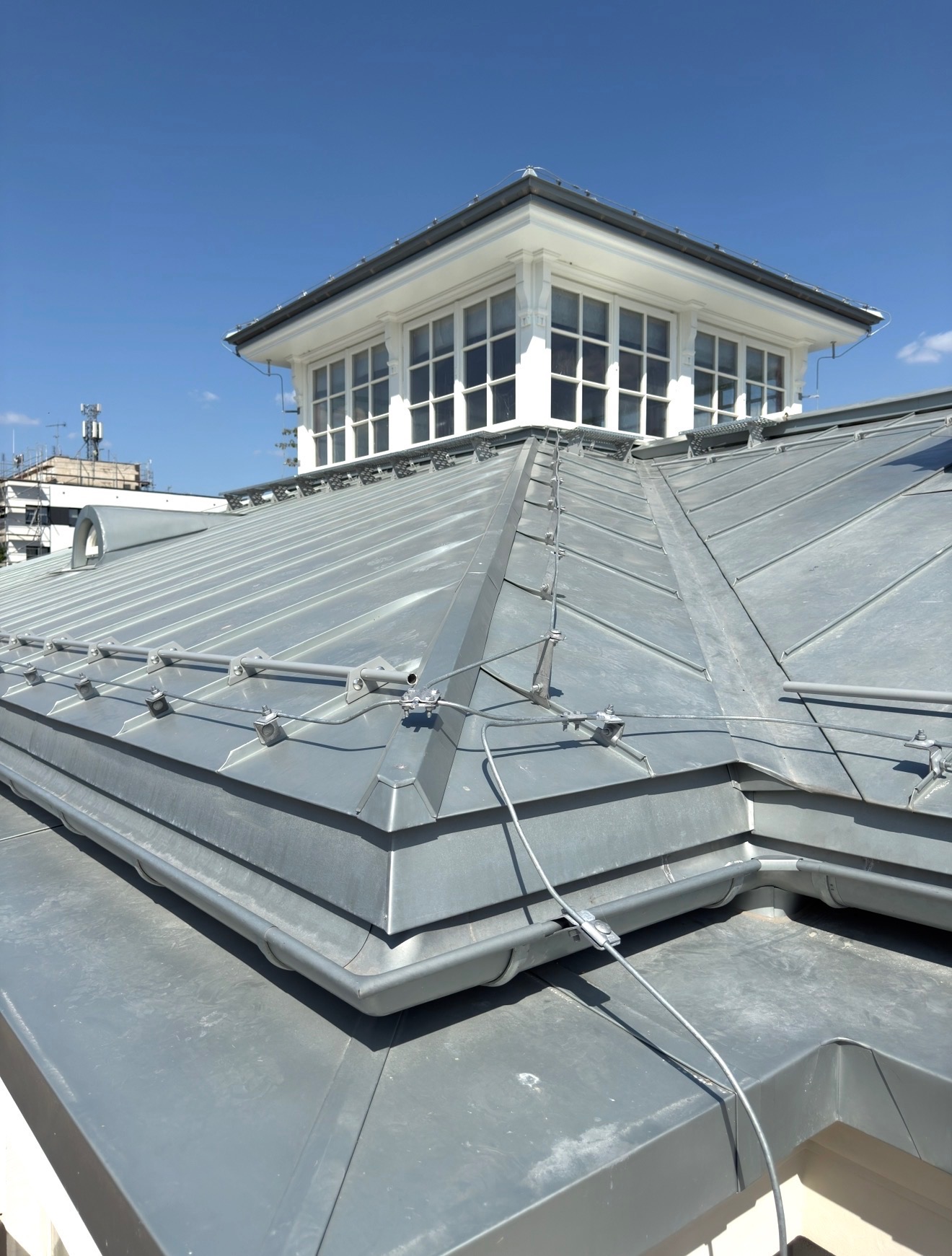 Metalowy dach z odgromem i lukarną w Warszawie, wykonany z blachy na rąbek stojący. Widok z dołu, błękitne niebo w tle.