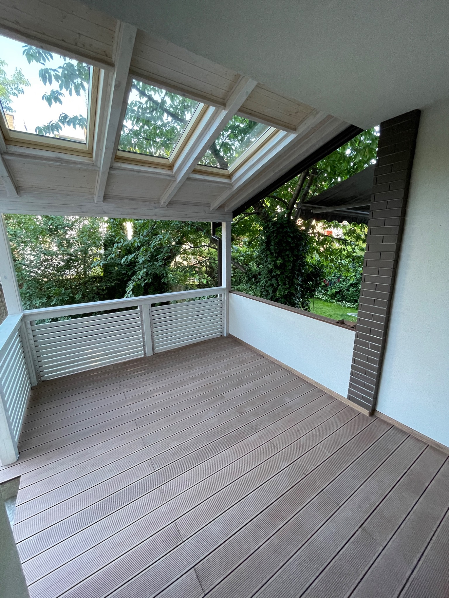 Zadaszony taras z drewnianą podłogą kompozytową, białymi balustradami i oknami dachowymi z widokiem na zieleń.