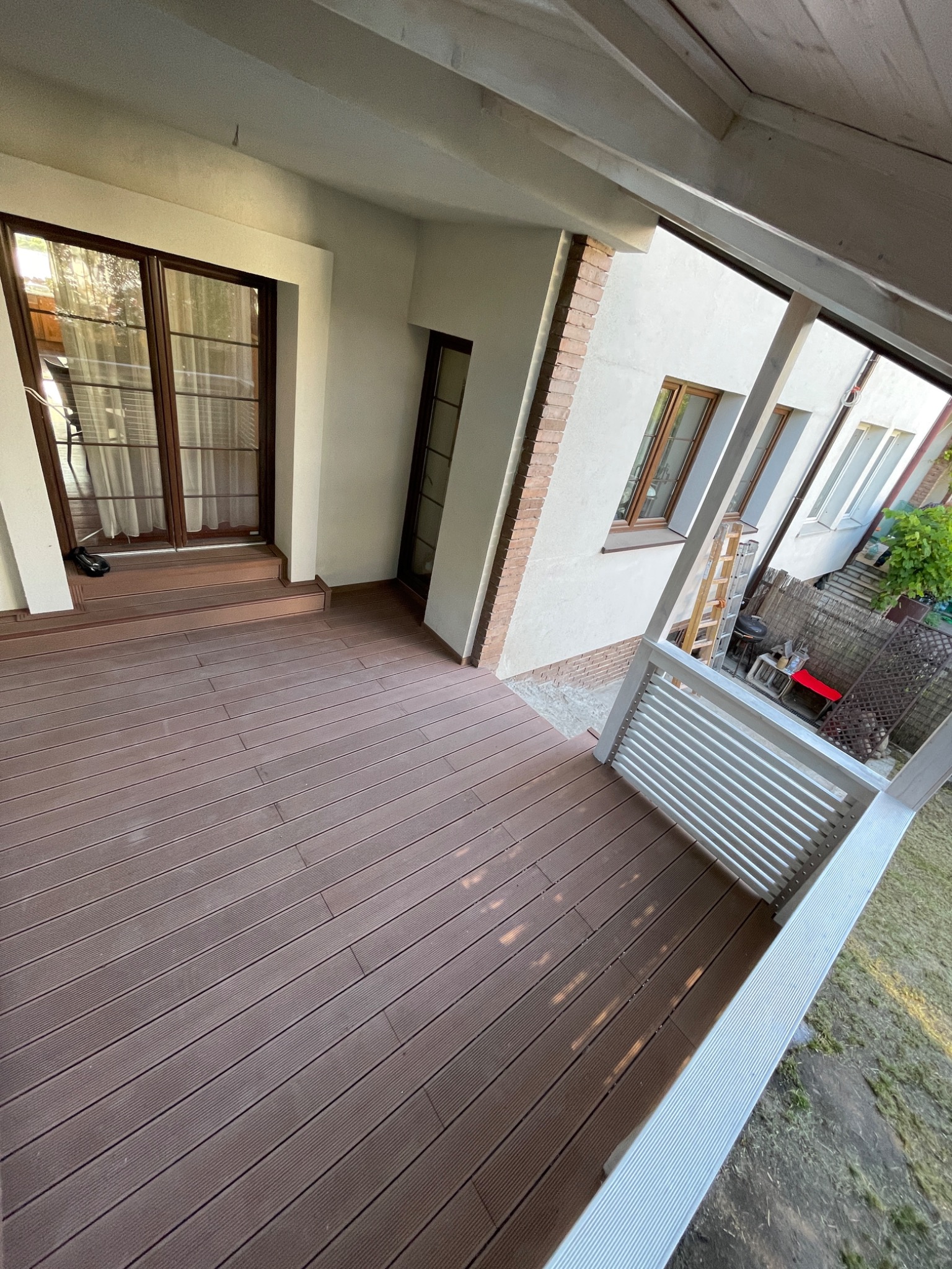 Zadaszony taras z kompozytowych desek w kolorze ciemnego brązu, biała balustrada, widok na okna budynku i drabinę opartą o ścianę.