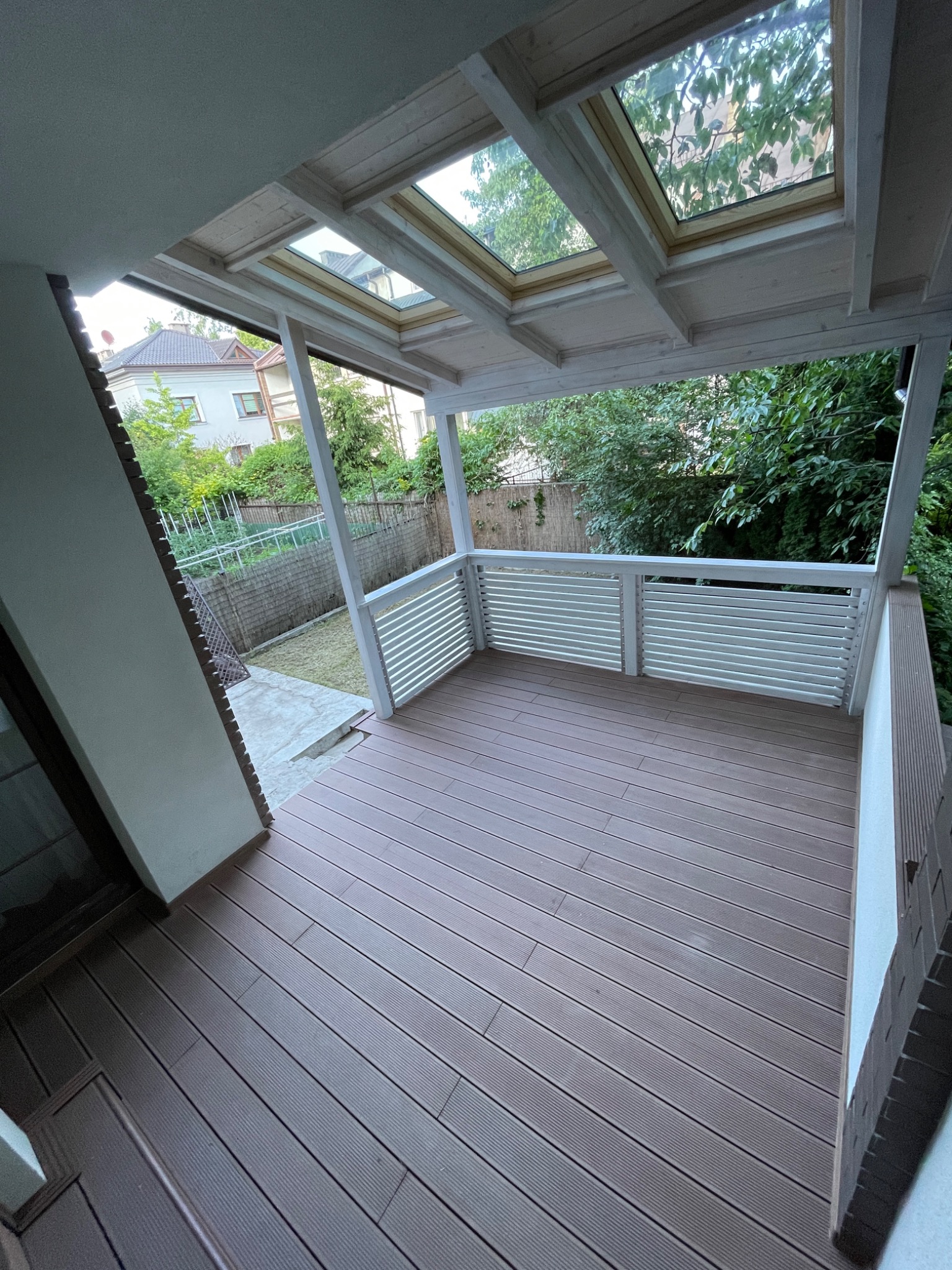 Zadaszony taras z brązową deską kompozytową i białymi balustradami, widok na ogród przez okna dachowe.