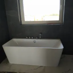 Nowoczesna, wolnostojąca wanna w łazience z ciemnymi, grafitowymi płytkami na ścianach i marmurową posadzką, widziana pod kątem, z oknem w tle.