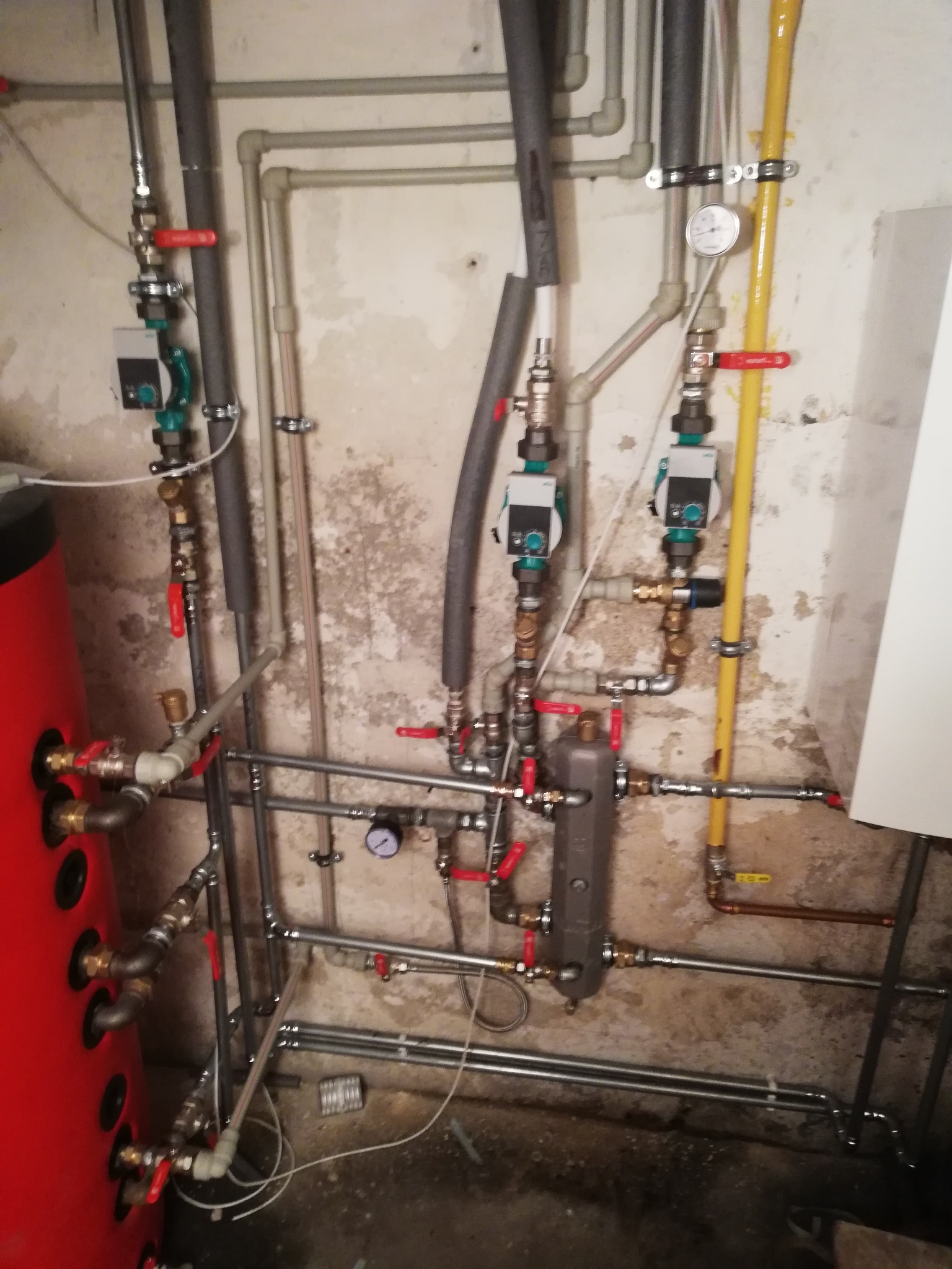 Złożona instalacja centralnego ogrzewania z czerwonym zasobnikiem ciepłej wody, widoczne rury, zawory, pompy obiegowe i manometry na tle surowej ściany piwnicy.