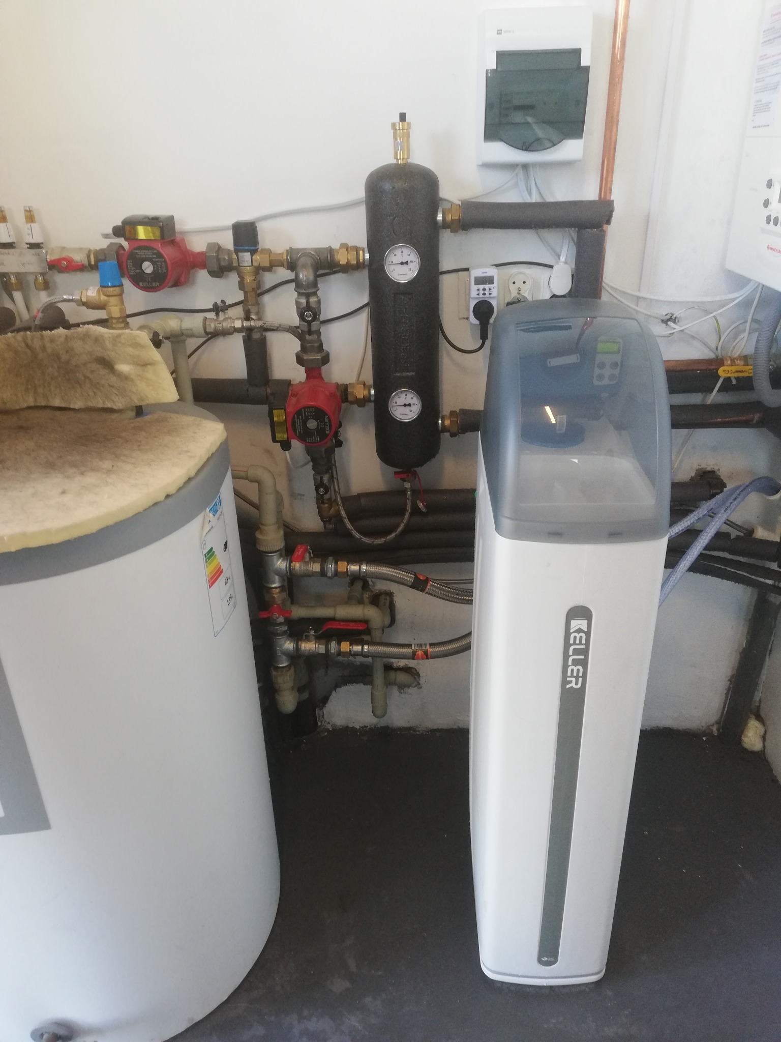 Centrala uzdatniania wody marki Keller obok zasobnika ciepłej wody z izolacją, widoczne rury, zawory, manometry i pompy obiegowe.
