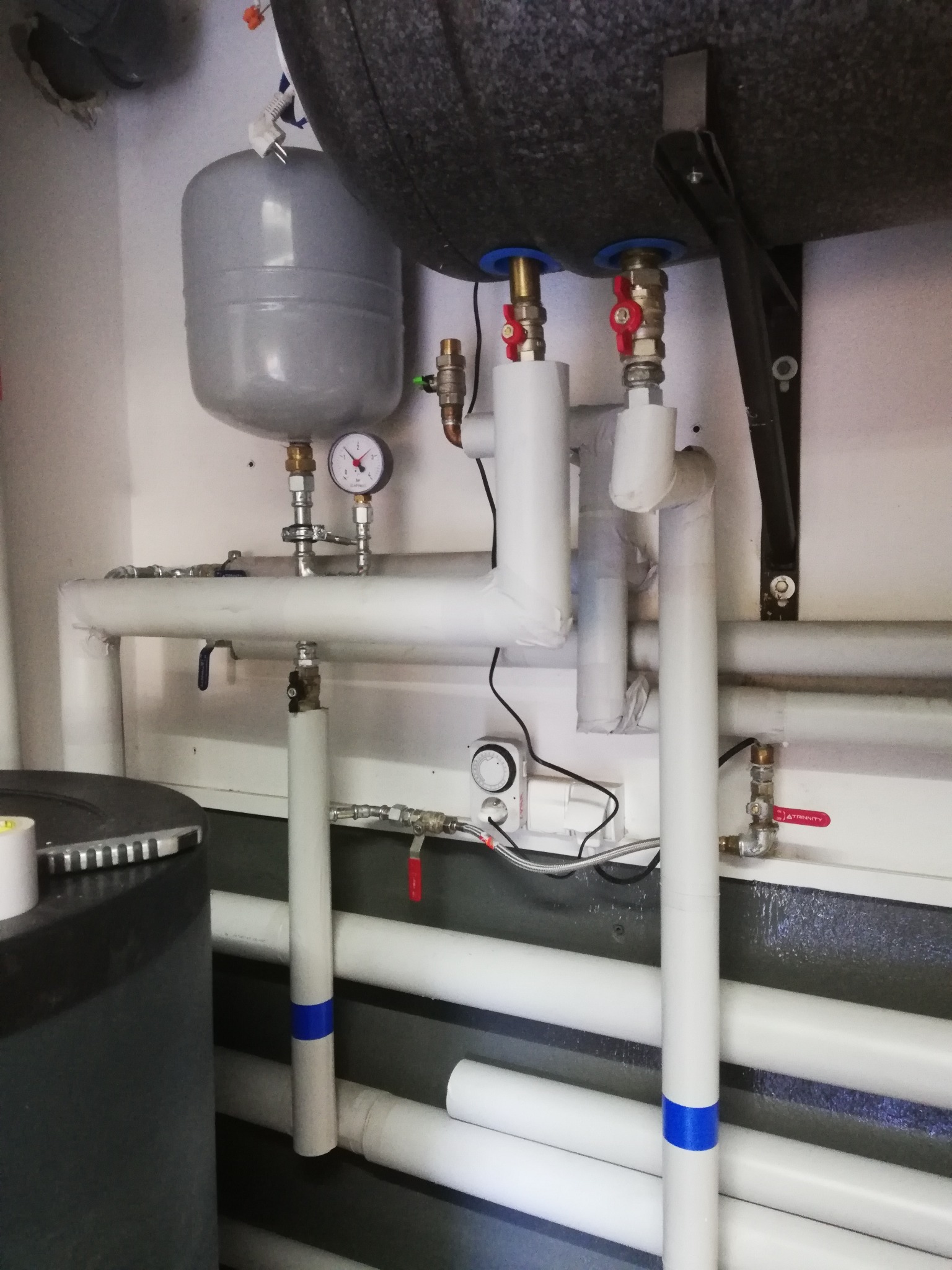 Zbiornik wyrównawczy i zasobnik ciepłej wody z zaworami, manometrem i białymi rurami instalacji grzewczej w kotłowni, widoczne elementy sterujące i izolacja.