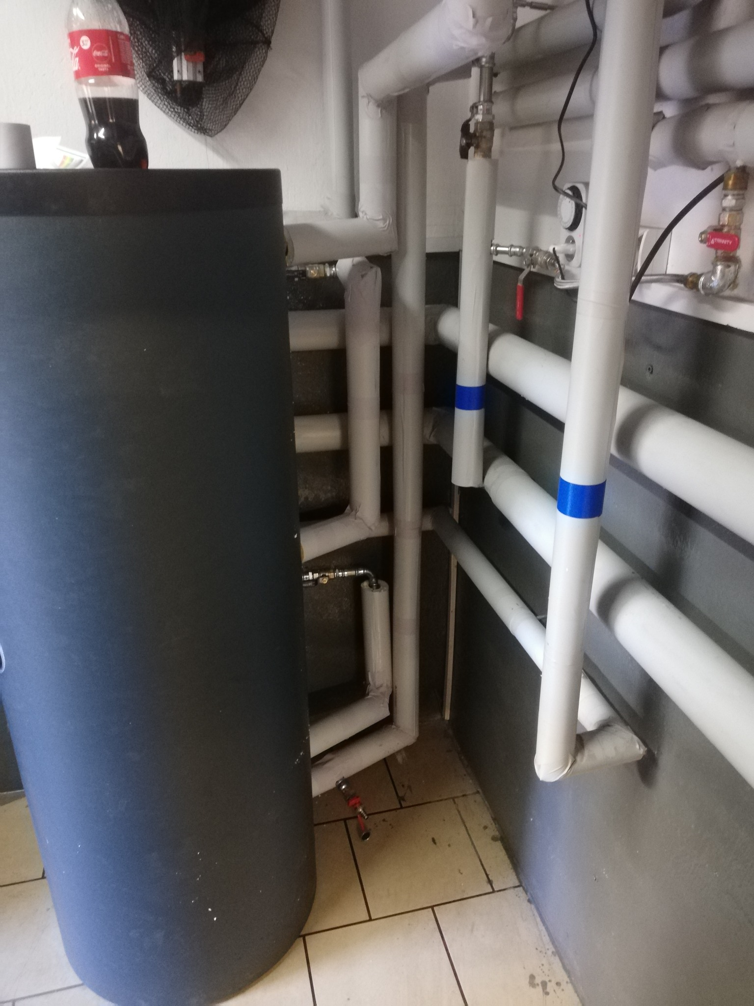 Ciemnoszary zasobnik ciepłej wody z widocznymi białymi rurami instalacji grzewczej, niektóre z niebieskimi oznaczeniami, w pomieszczeniu z wykafelkowaną podłogą i butelką Coca-Coli na górze zbiornika.