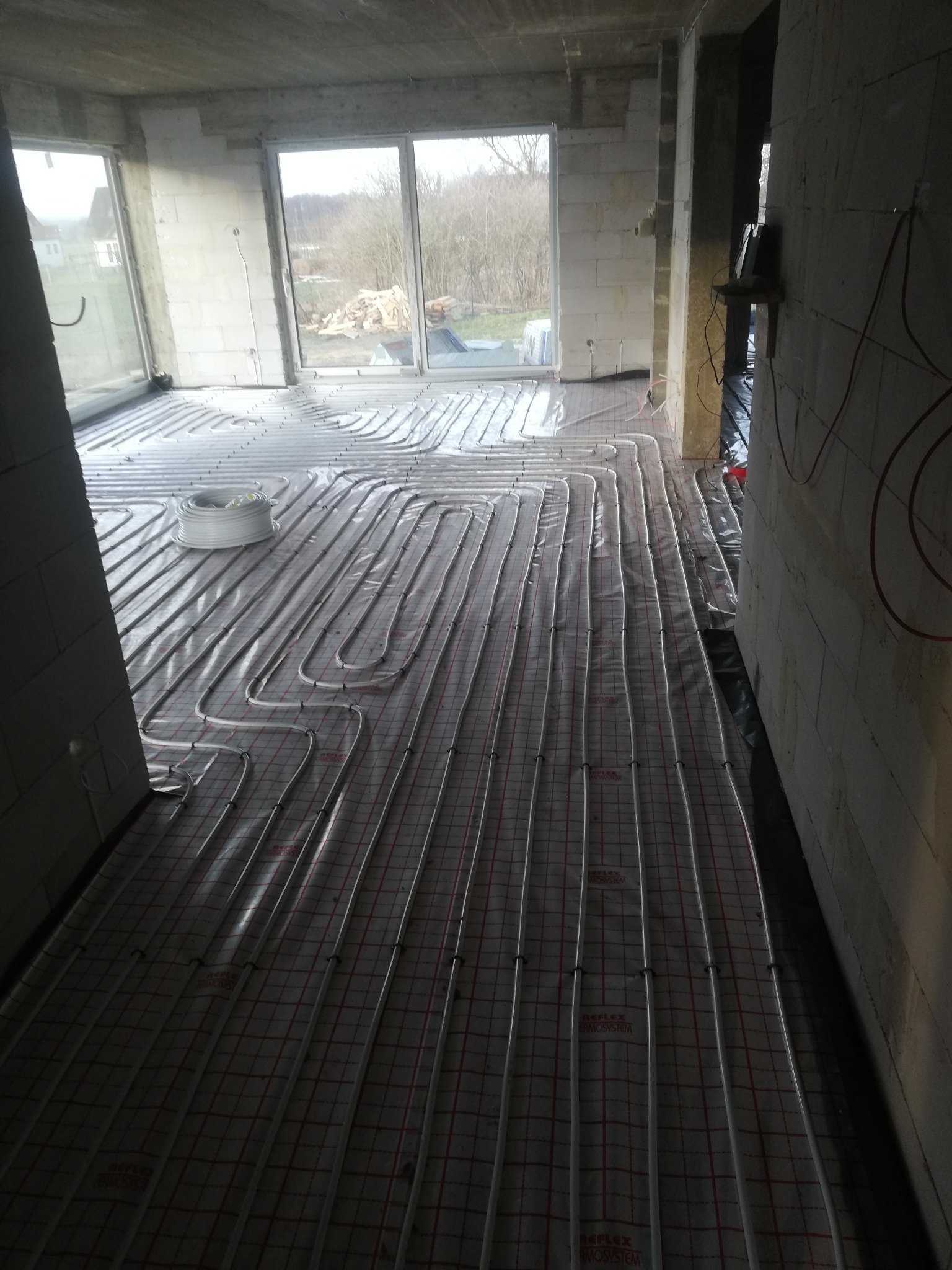 Montaż ogrzewania podłogowego w surowym stanie, rury grzewcze ułożone na folii z siatką, widoczna rolka rur, ściany z pustaków, okna z widokiem na drzewa.