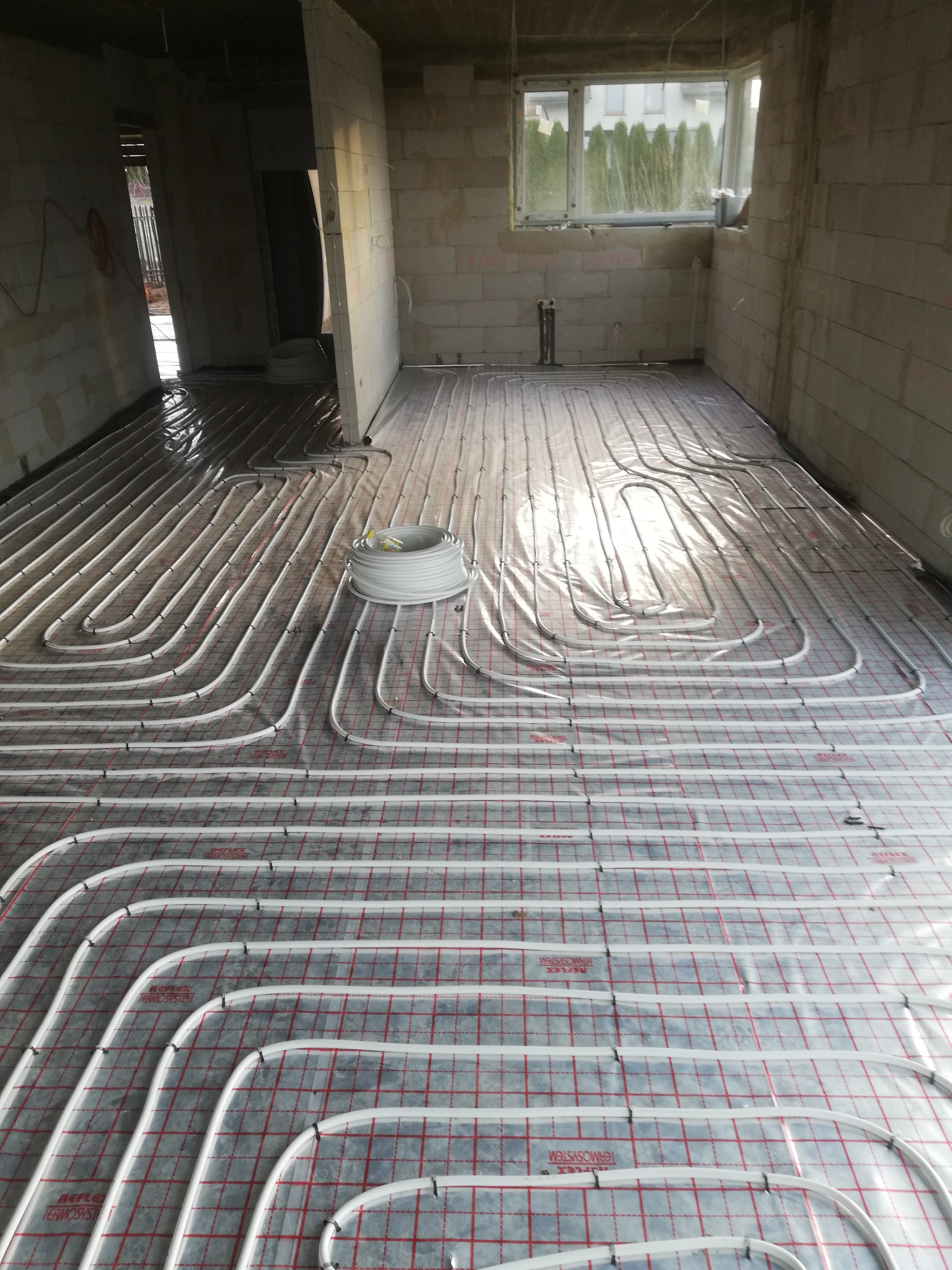 Rozłożone rury ogrzewania podłogowego na folii z siatką w surowym wnętrzu budynku, widoczny zwój rur i fragment okna.