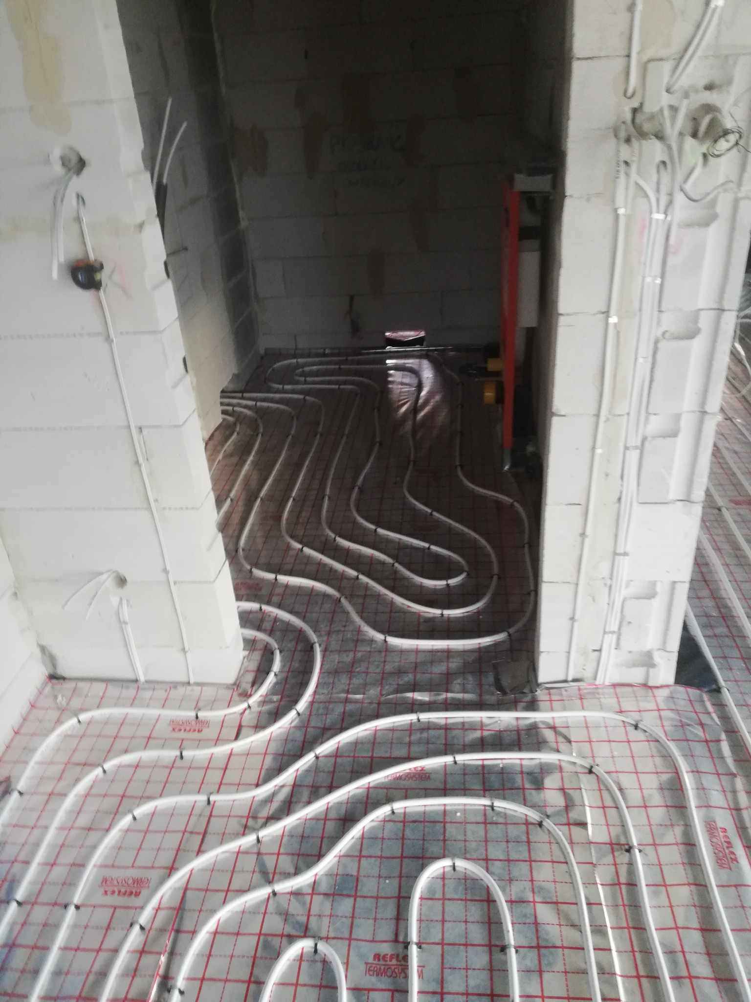 Rozłożone białe rury ogrzewania podłogowego na folii z czerwoną siatką w surowym wnętrzu budynku z białymi ścianami z betonu komórkowego.