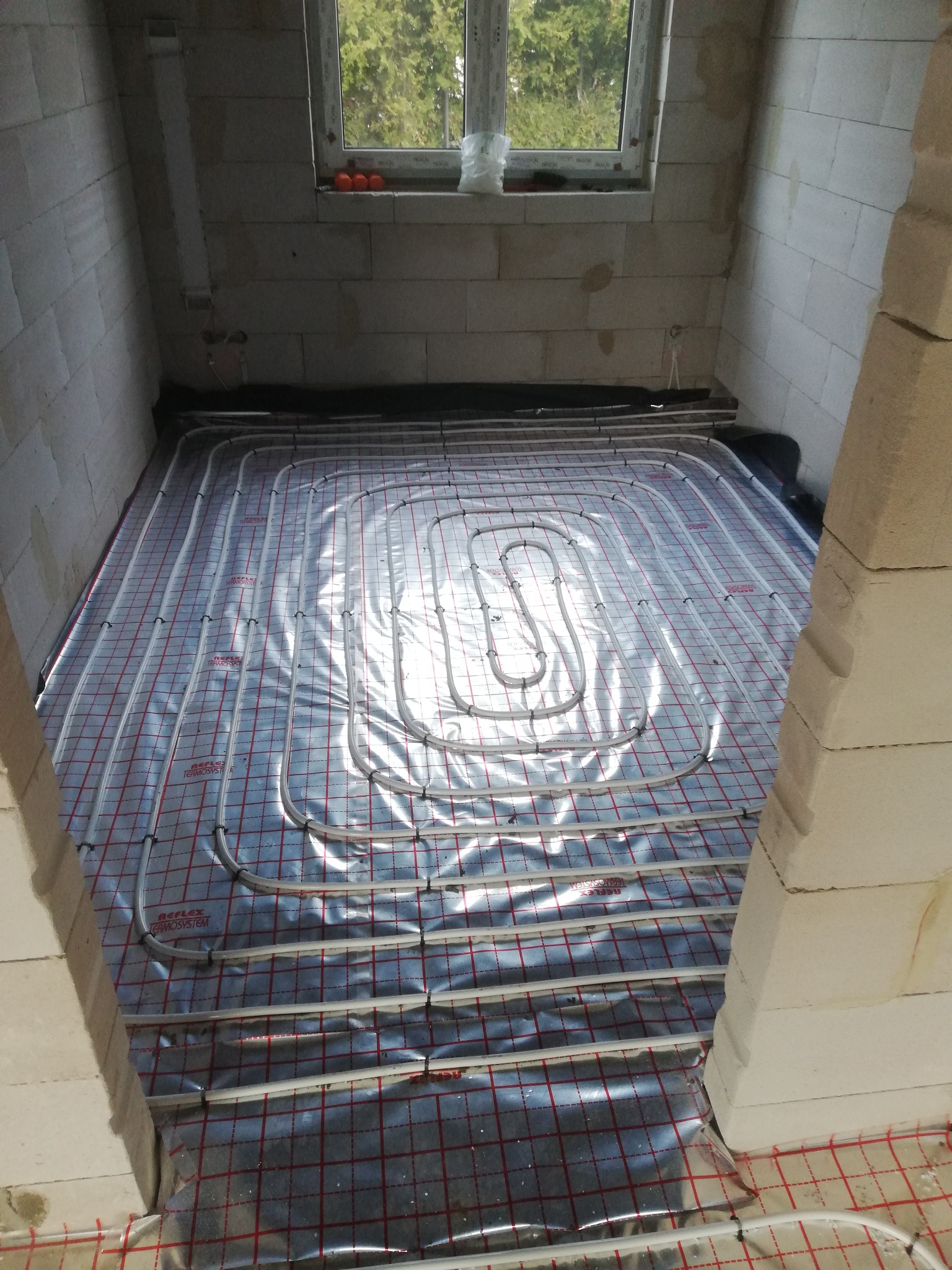 Rozłożona instalacja ogrzewania podłogowego na folii z czerwoną siatką w surowym pomieszczeniu z oknem. Widoczne białe rurki przymocowane do podłoża czarnymi klipsami. W tle widoczne trzy...