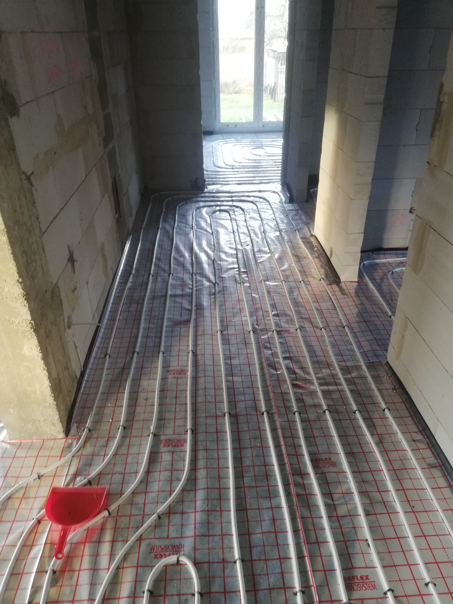 Rury ogrzewania podłogowego ułożone na folii z czerwoną siatką w surowym wnętrzu budynku w trakcie budowy, widoczna czerwona szufelka.