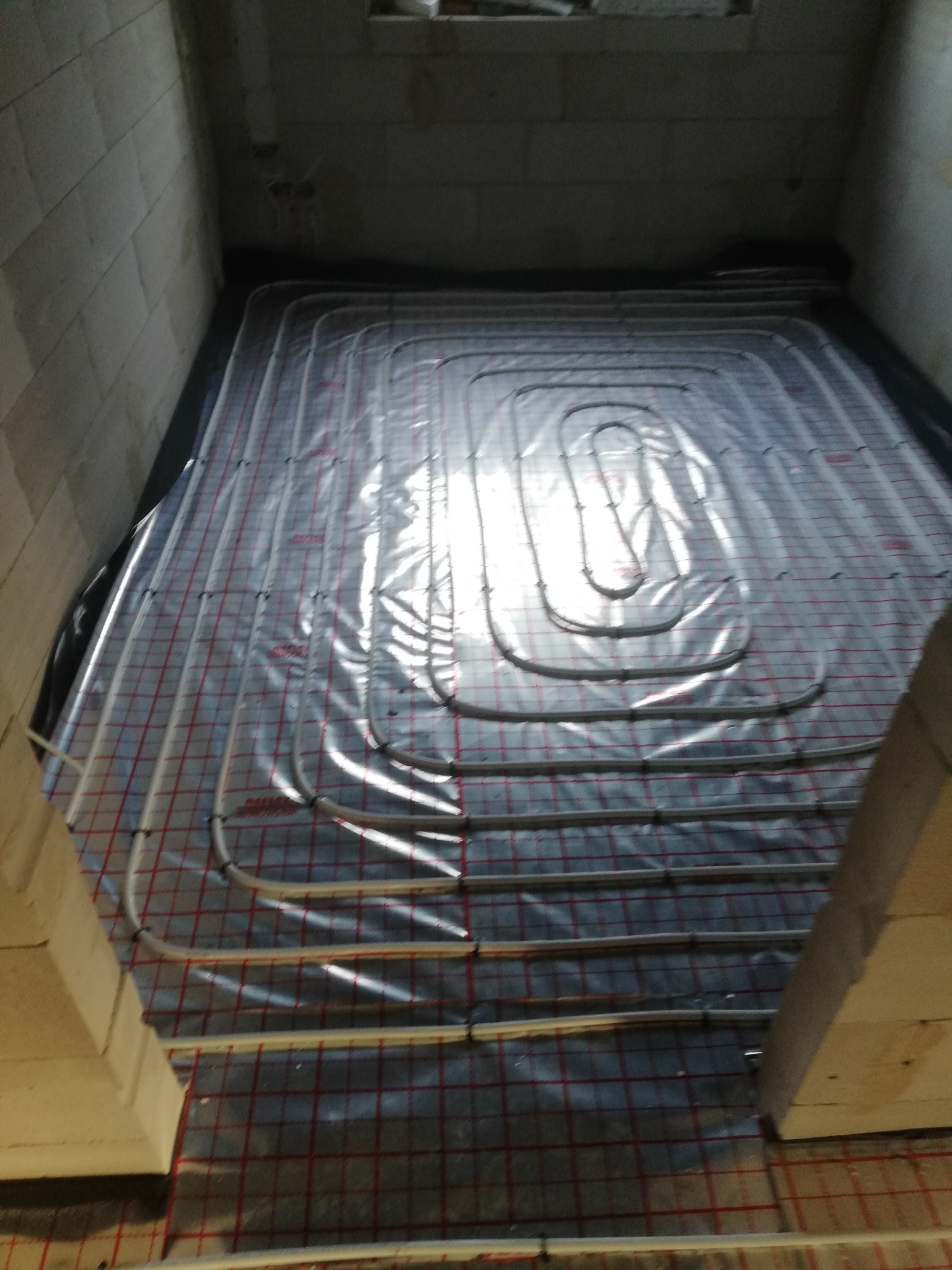 Rozłożona instalacja ogrzewania podłogowego na czarnej folii, z widoczną siatką z czerwonymi liniami, w surowym pomieszczeniu z betonowymi ścianami.