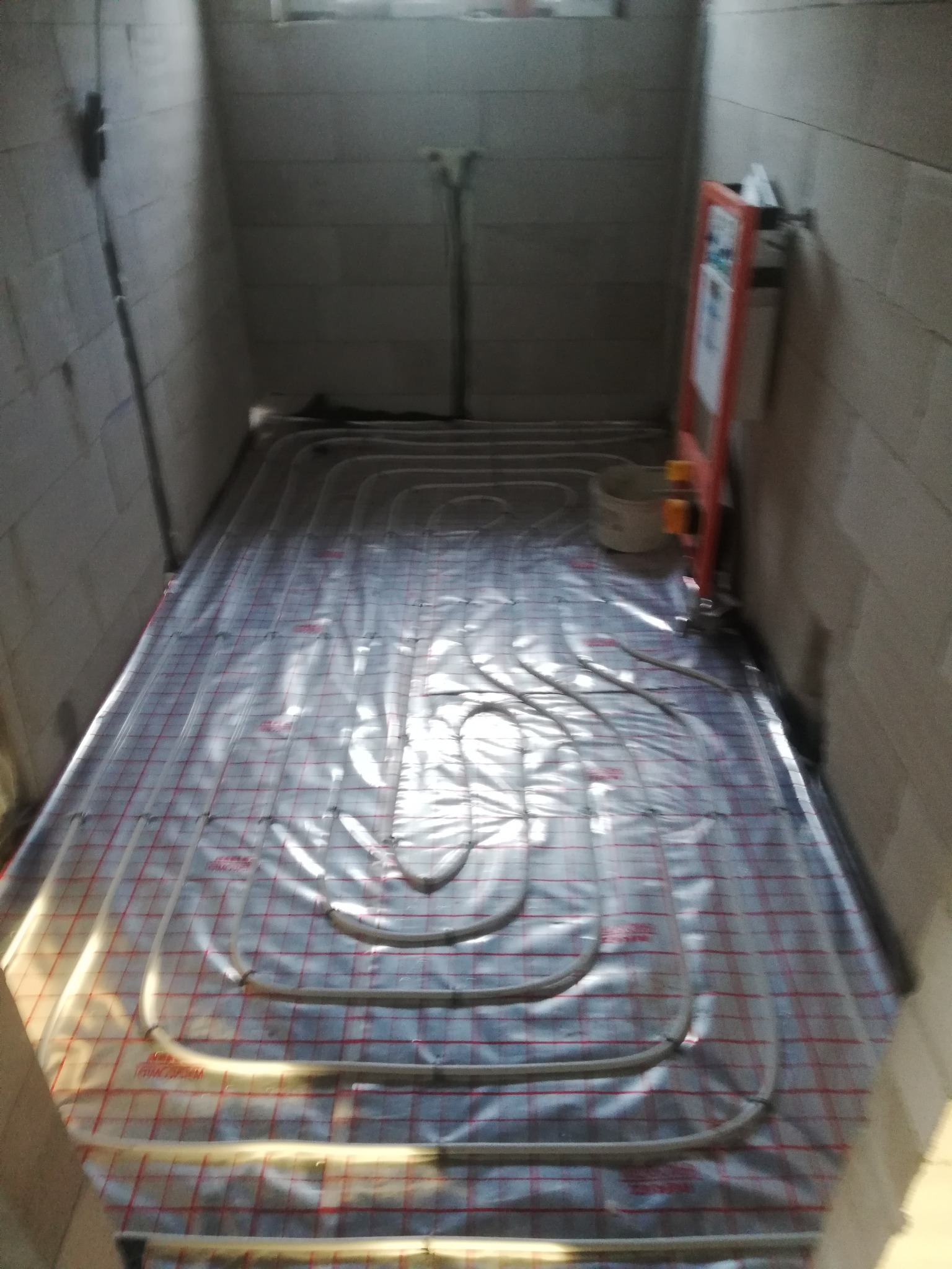 Rury ogrzewania podłogowego rozłożone na folii z siatką w surowym pomieszczeniu z bloczków betonowych przed wylewką.