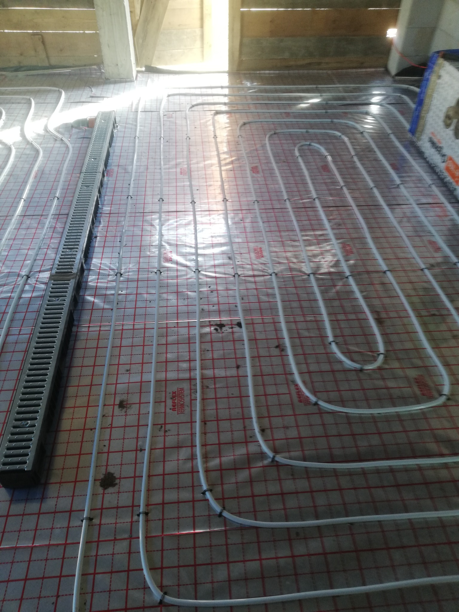 Rozłożona instalacja ogrzewania podłogowego z rurkami na folii z siatką, widoczny metalowy odpływ liniowy, surowy strop w tle.