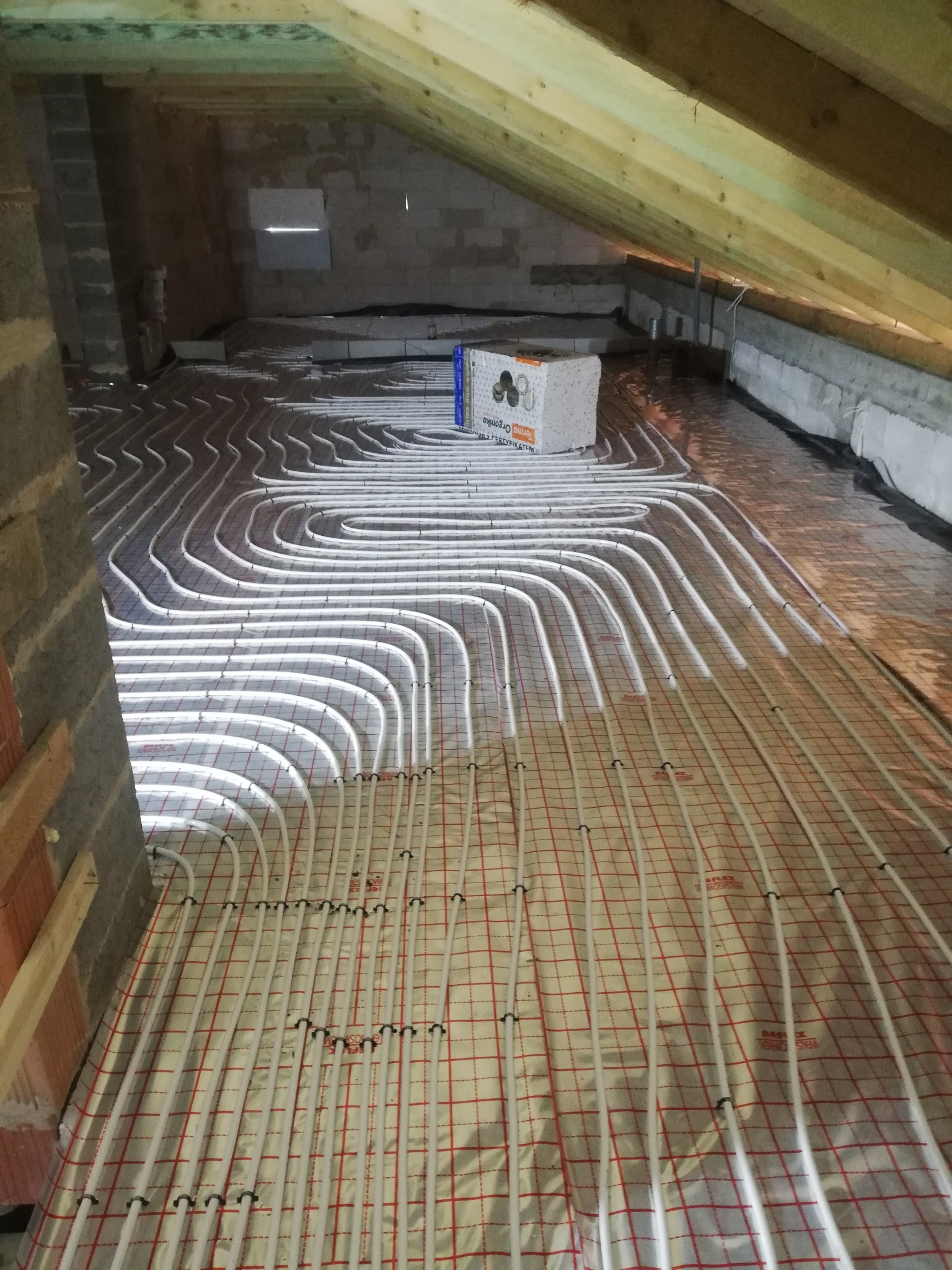 Białe rury ogrzewania podłogowego ułożone na folii refleksyjnej z czerwoną siatką w surowym pomieszczeniu na poddaszu, widoczny bloczek z betonu komórkowego i drewniana konstrukcja dachu.