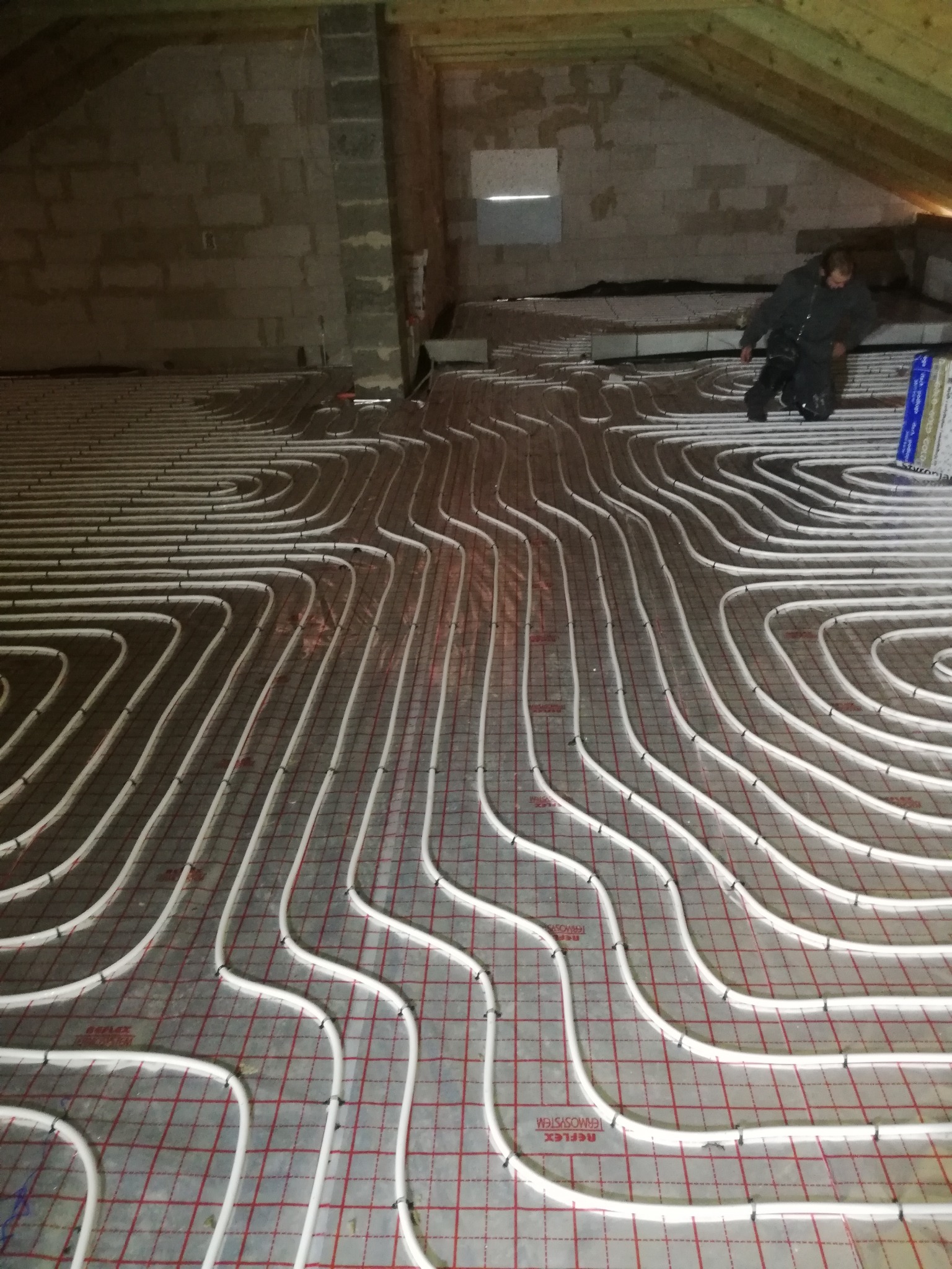 Białe rury ogrzewania podłogowego ułożone na folii z czerwoną siatką, widoczna osoba w roboczym ubraniu w trakcie montażu, surowy strop z betonu komórkowego w tle.