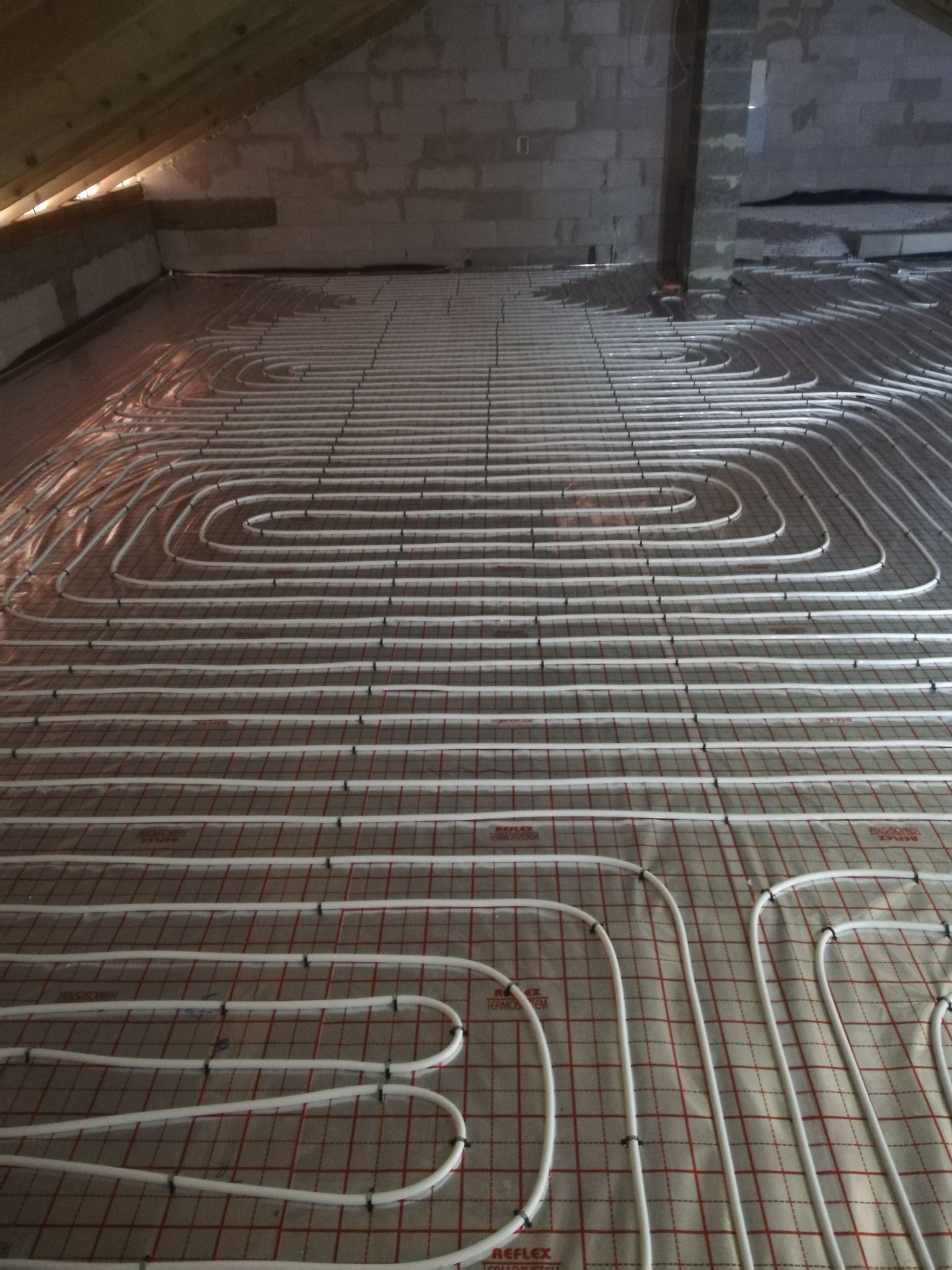 Rozłożone rury ogrzewania podłogowego na izolacji z siatką w surowym wnętrzu poddasza z widoczną konstrukcją dachu.