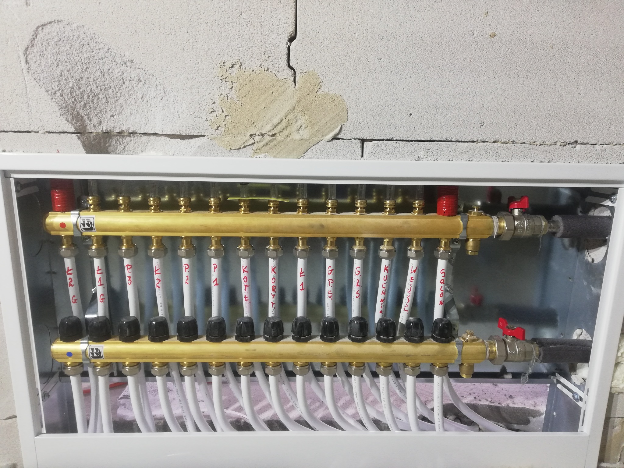Rozdzielacz podłogowy z rurami oznaczonymi nazwami pomieszczeń, zamontowany w białej skrzynce na tle surowej ściany z betonu komórkowego z widocznymi śladami po łataniu.