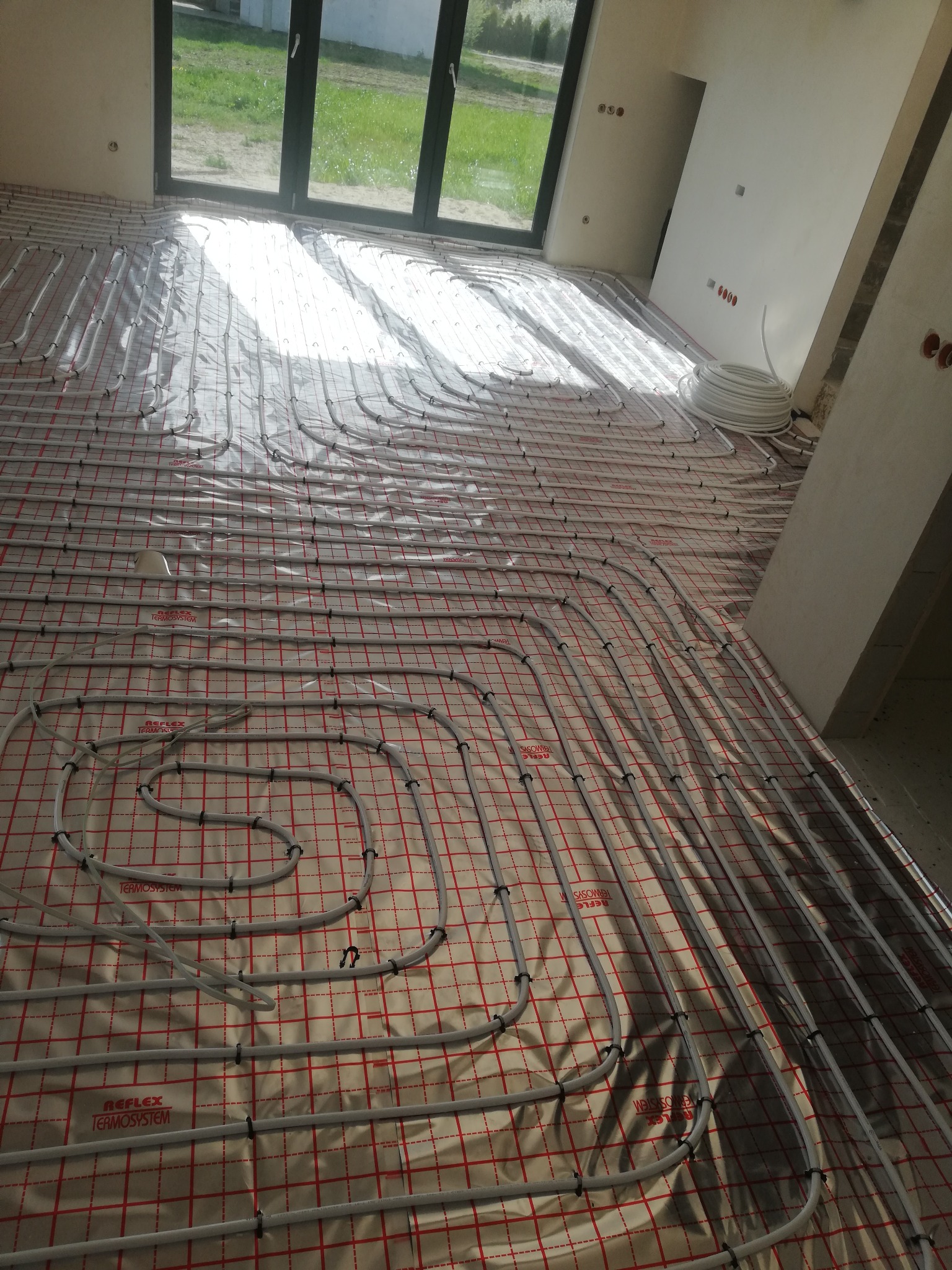 Białe rury ogrzewania podłogowego ułożone w pętle na folii z czerwoną kratką w jasnym pomieszczeniu z dużym oknem balkonowym i widocznym fragmentem zwiniętej rury.