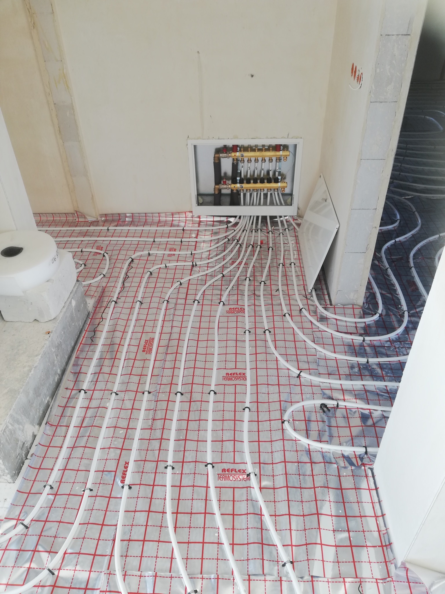 Rozłożone białe rury ogrzewania podłogowego na folii z czerwoną kratką, widoczny rozdzielacz w ścianie, surowy stan wykończenia wnętrza.
