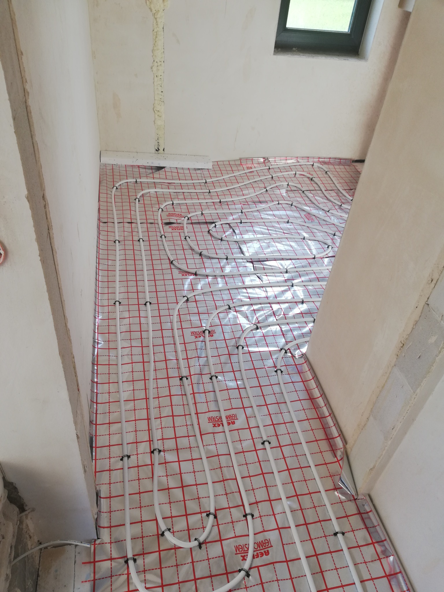 Białe rurki ogrzewania podłogowego, zamocowane czarnymi uchwytami, ułożone na srebrnej folii z czerwoną kratką w surowym wnętrzu podczas montażu.