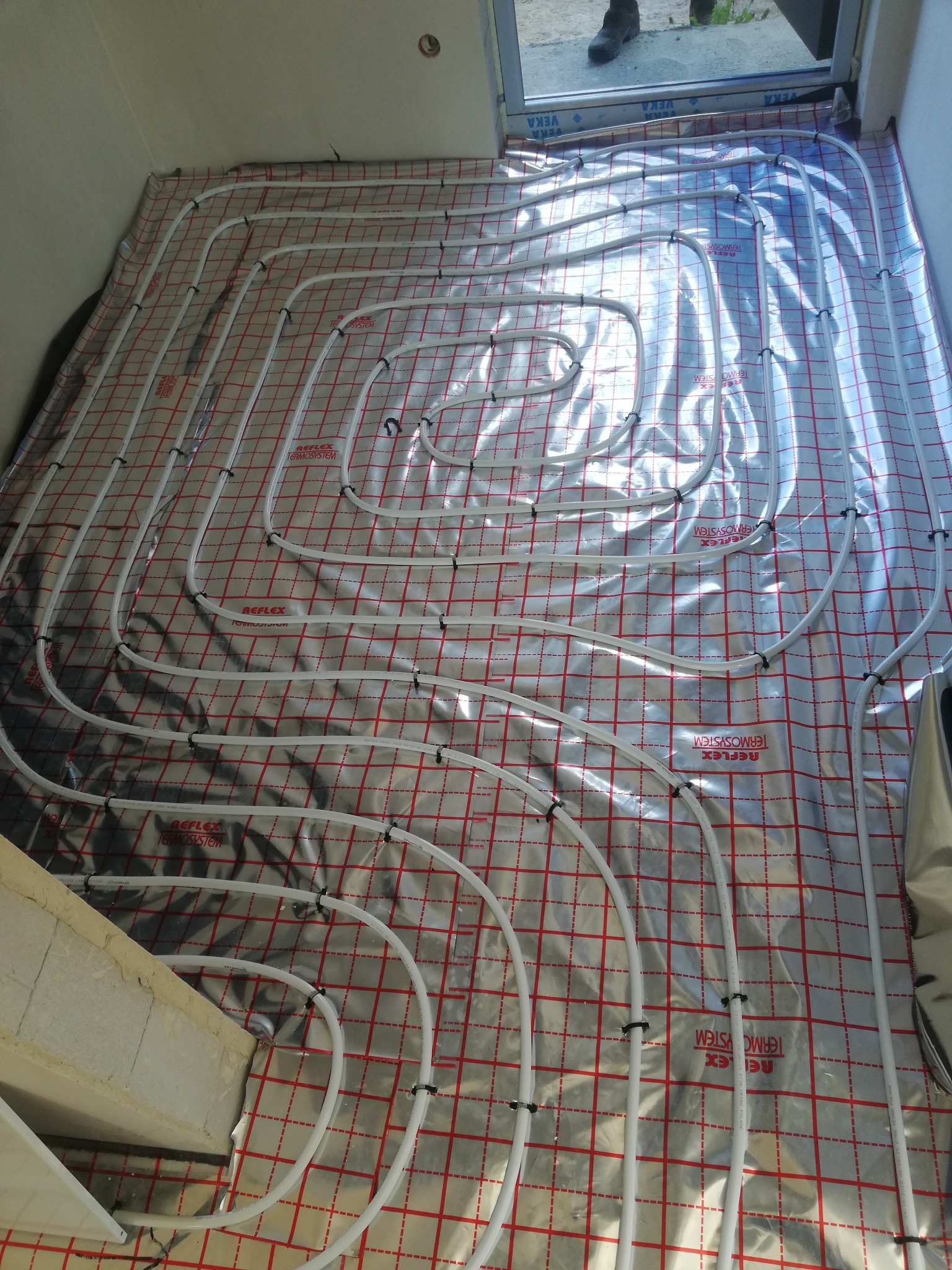 Rozłożone białe rurki ogrzewania podłogowego na srebrnej folii z czerwoną kratką, widoczne czarne spinki mocujące, fragment ściany i okna w tle.