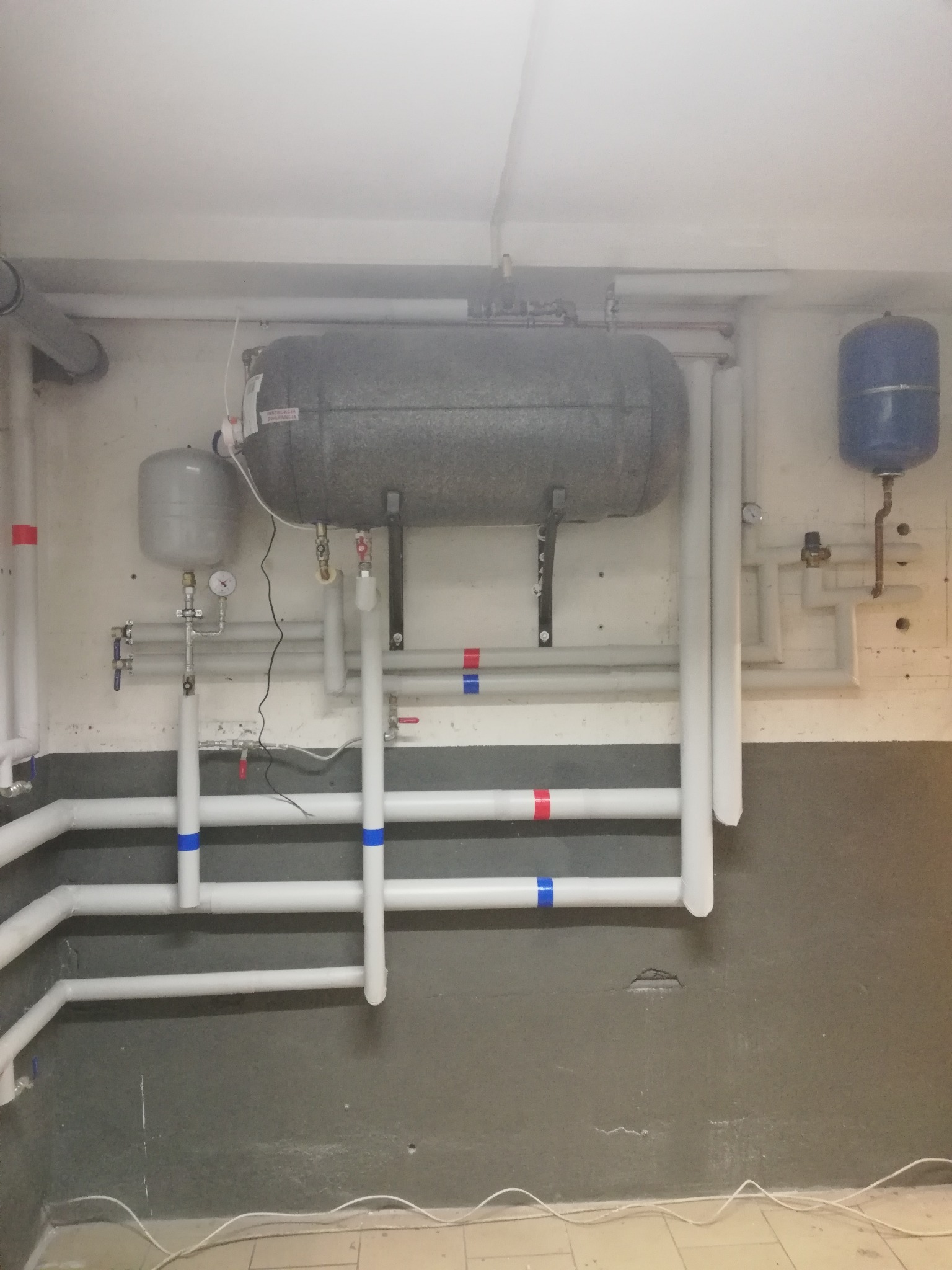 Piwnica z zainstalowanym poziomym zbiornikiem akumulacyjnym ciepła, białymi rurami z kolorowymi oznaczeniami, manometrem i zaworami.