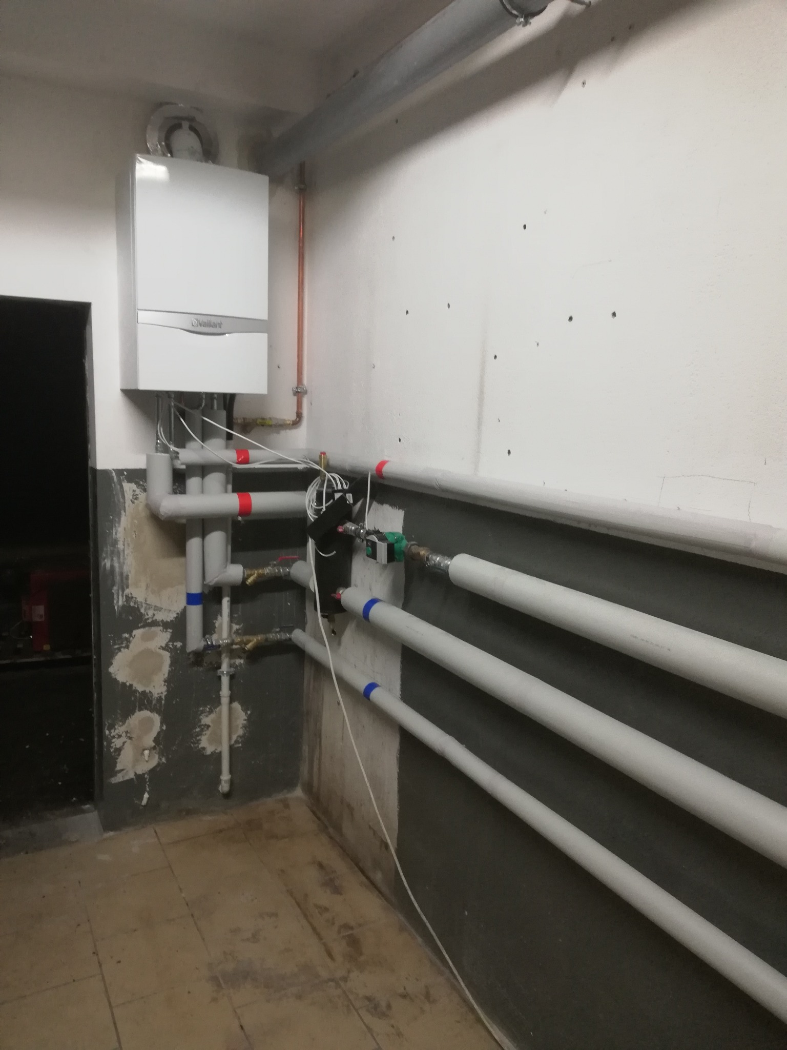 Biały wiszący piec gazowy marki Vaillant z szarymi rurami instalacji centralnego ogrzewania w surowym pomieszczeniu z częściowo odrapanymi ścianami i brudną podłogą z płytek.