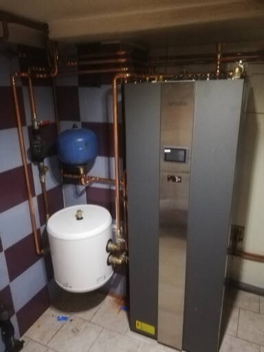Zainstalowany nowoczesny piec grzewczy z zasobnikiem ciepłej wody i niebieskim zbiornikiem wyrównawczym, połączony miedzianymi rurami w pomieszczeniu z kafelkami w kratę.