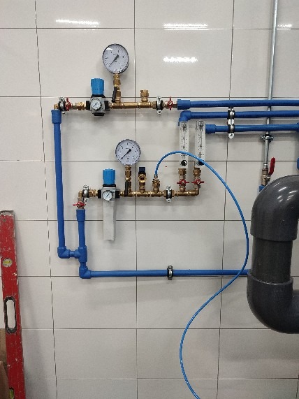 Kompleksowy system rur wodnych i gazowych z manometrami i filtrami, zamontowany na kafelkowanej ścianie, widoczny poziomica z boku.